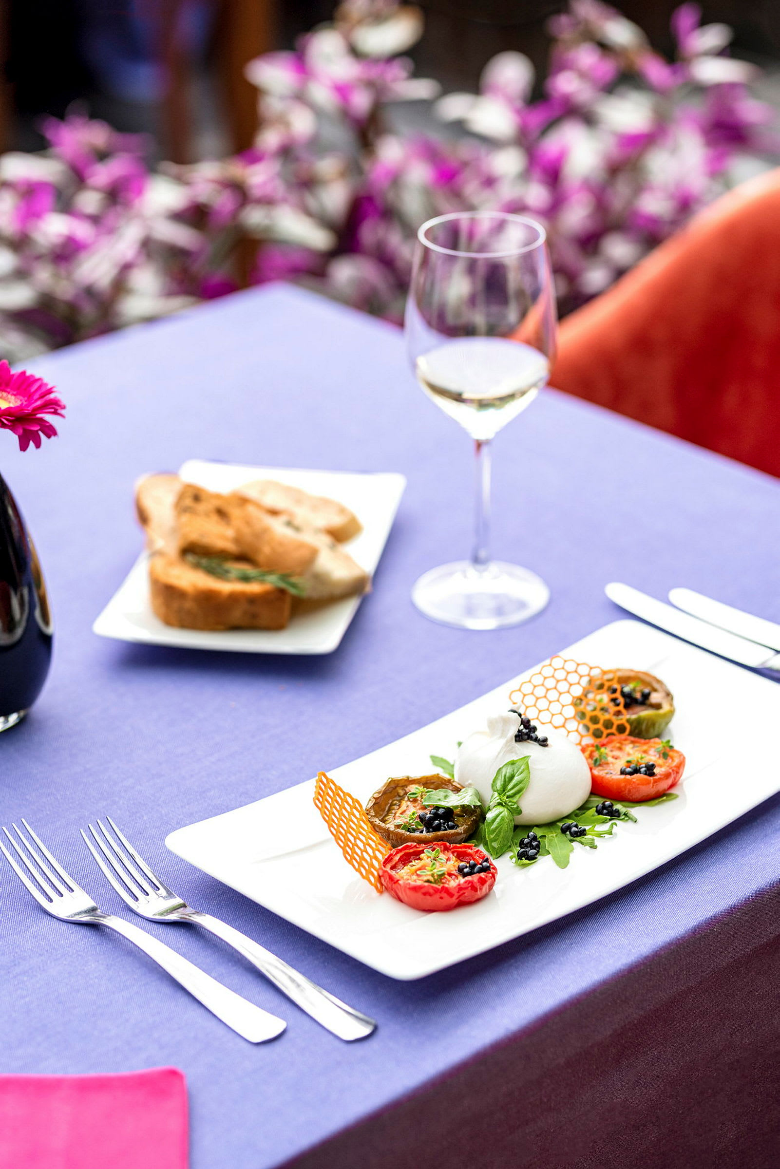 En tallerken med forrett som inneholder tomater, burrata og dekorasjoner, med et vinglass og en bolle med brød på en duk. (AI-generert tekst)