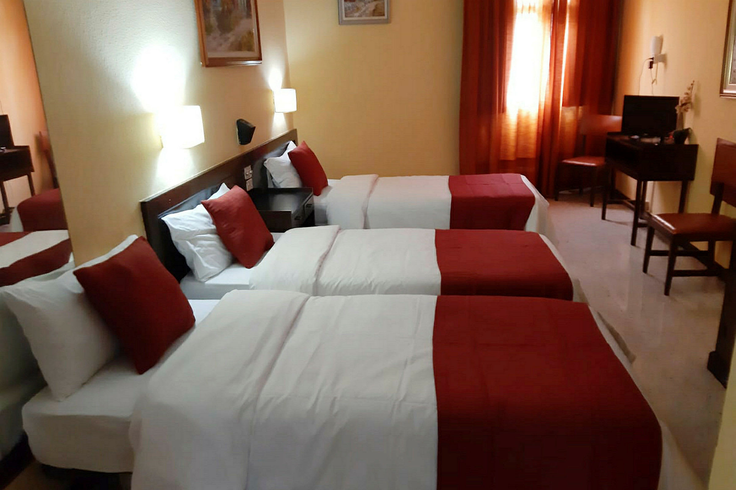 Et hotellrom med tre enkeltsenger, hvite laken og røde sengetepper, med en nattbord og belysning på veggen. (AI-generert tekst)
