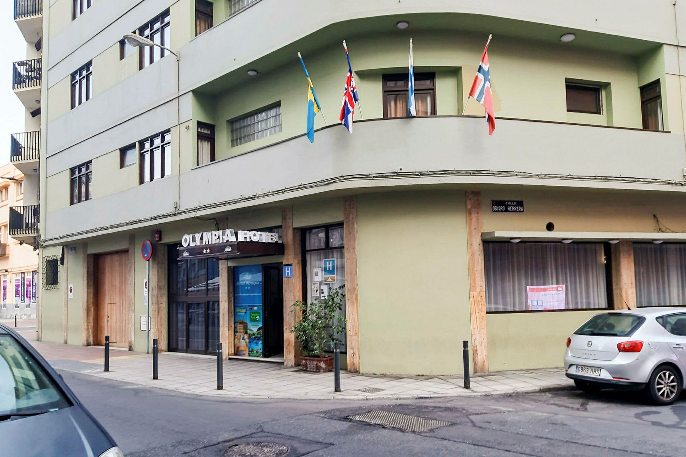 En grønn bygning med teksten 'Olympia Hotel' over inngangen. Flere flagg er satt opp på en balkong. (AI-generert tekst)