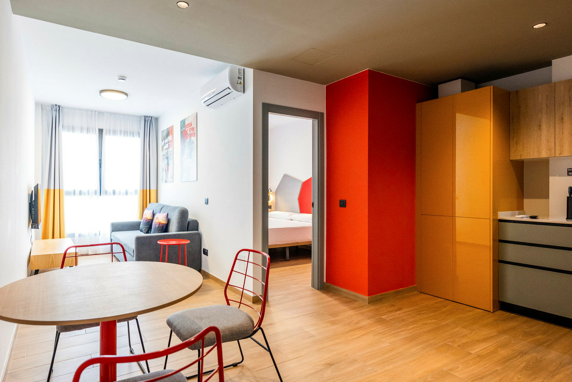Et moderne innredet hotellrom med et bord og stoler i forgrunnen, en sittegruppe med sofa og et soverom synlig gjennom en døråpning. Kjøkkenområdet har skap og arbeidsflater. (AI-generert tekst)