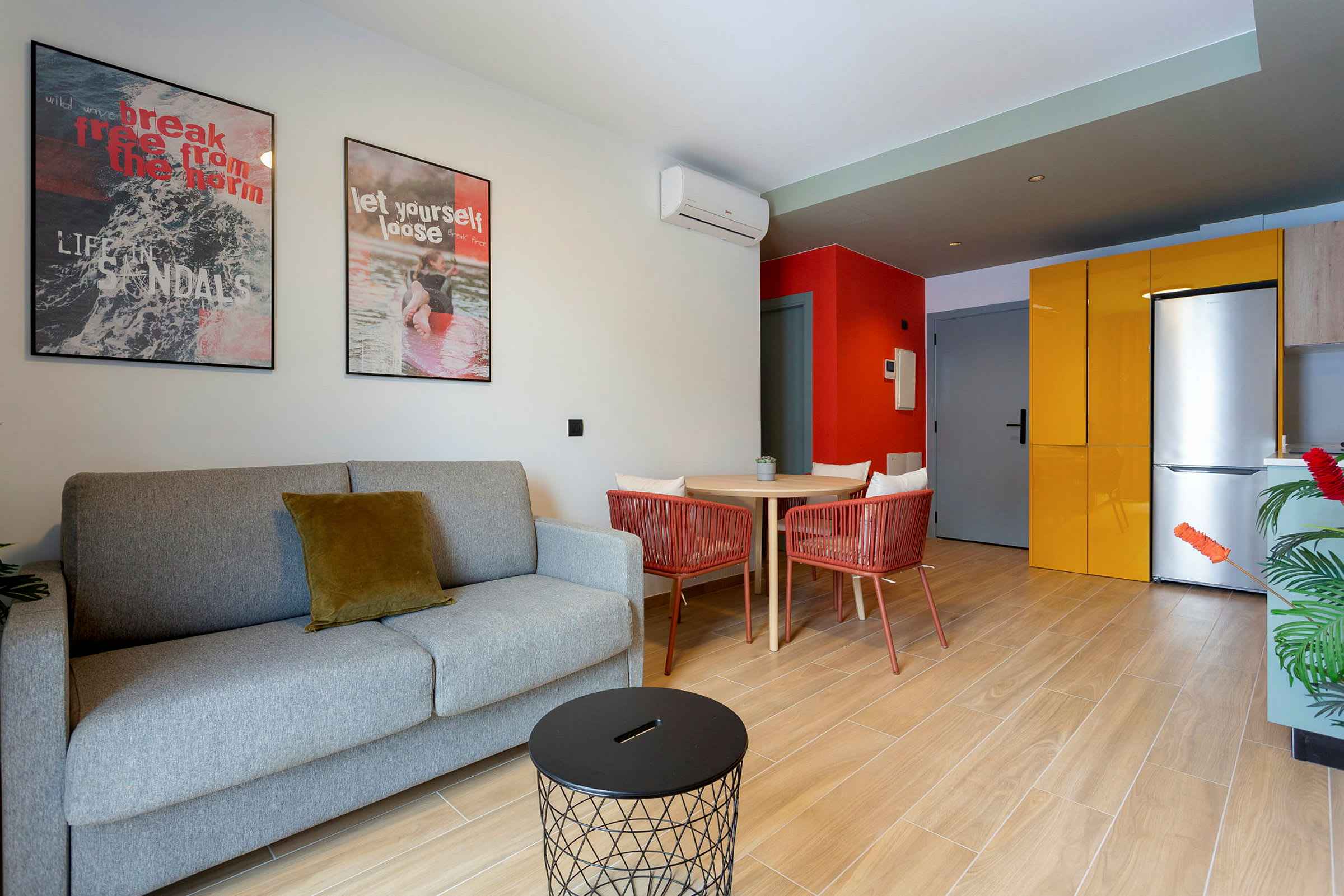 Et moderne stue med en grå sofa og et lite svart bord. På veggen er det to fargerike malerier. Til høyre sees et kjøkken med gul kjøkkeninnredning og et kjøleskap. (AI-generert tekst)