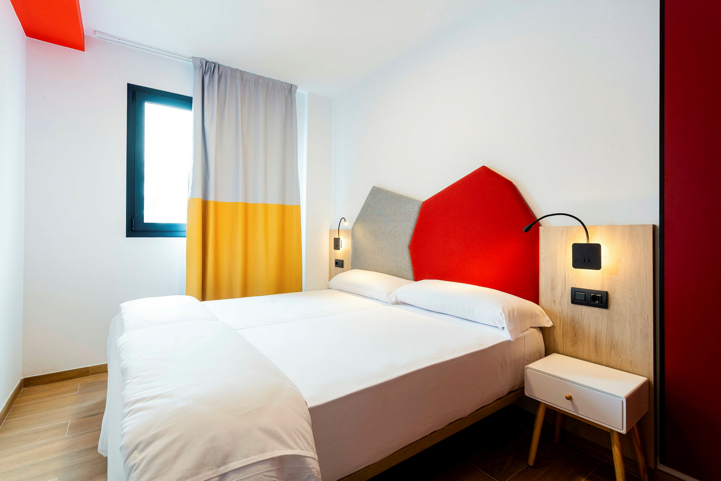 Et hotellrom med en dobbeltseng, fargerike veggdekorasjoner og et vindu med gardin. (AI-generert tekst)