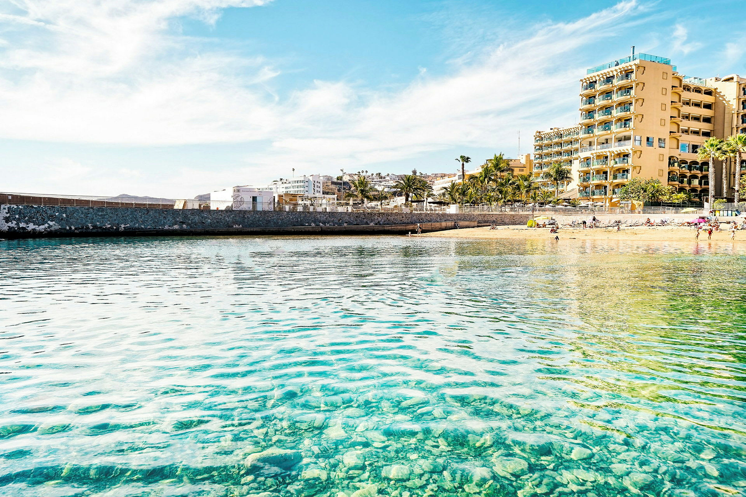 Strand med en steinmur og hotell i bakgrunnen. (AI-generert tekst)