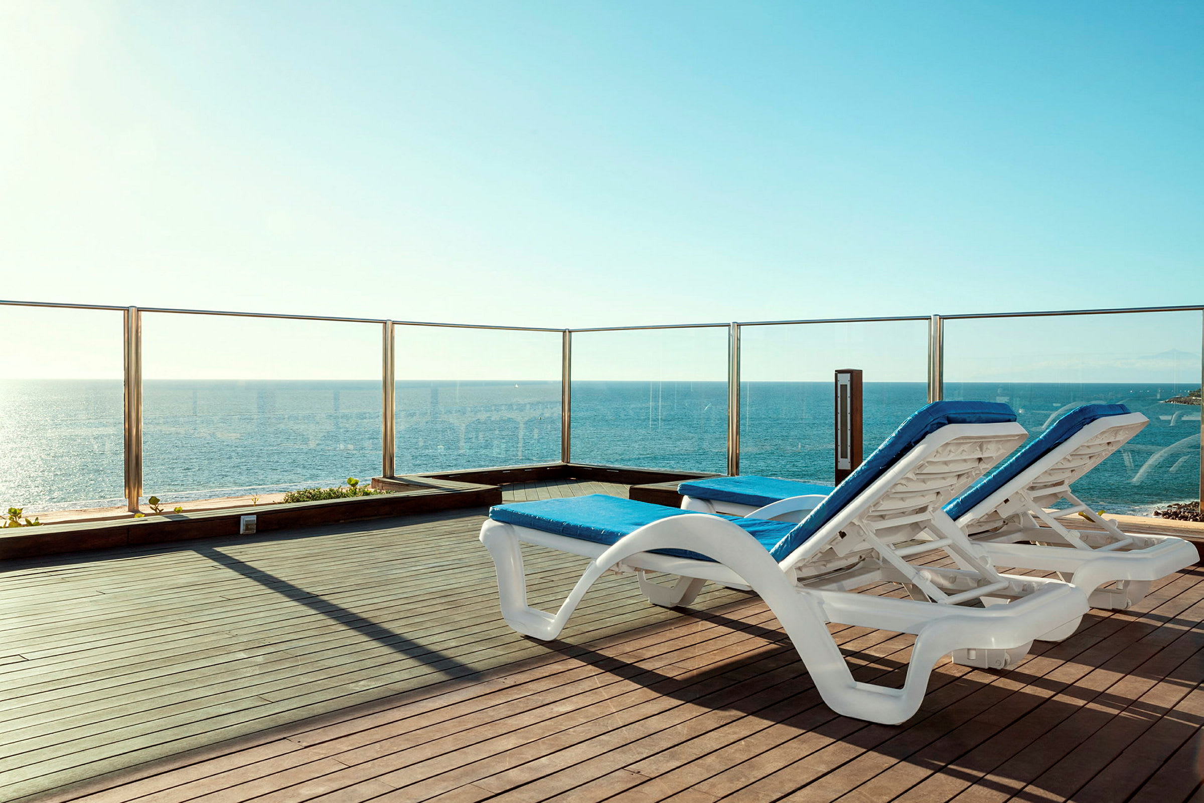 To solsenger med blå puter står på en terrasse med glassrekkverk og utsikt over havet. (AI-generert tekst)