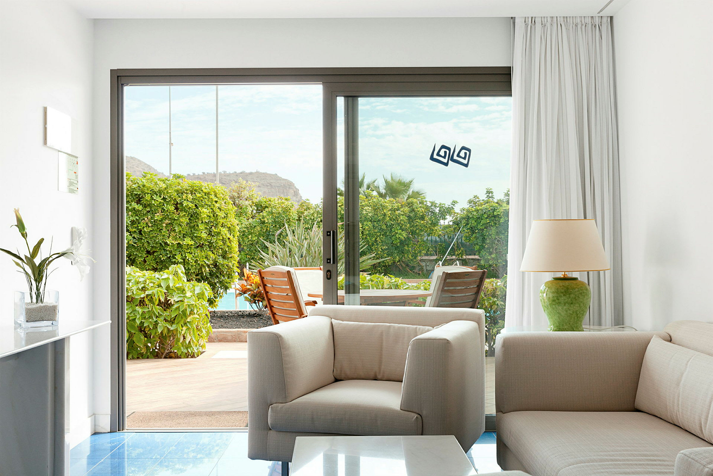 Bilde av et rom med lys innredning, en lenestol og sofa, med en glassdør som fører ut til en terrasse med grøntområder. (AI-generert tekst)