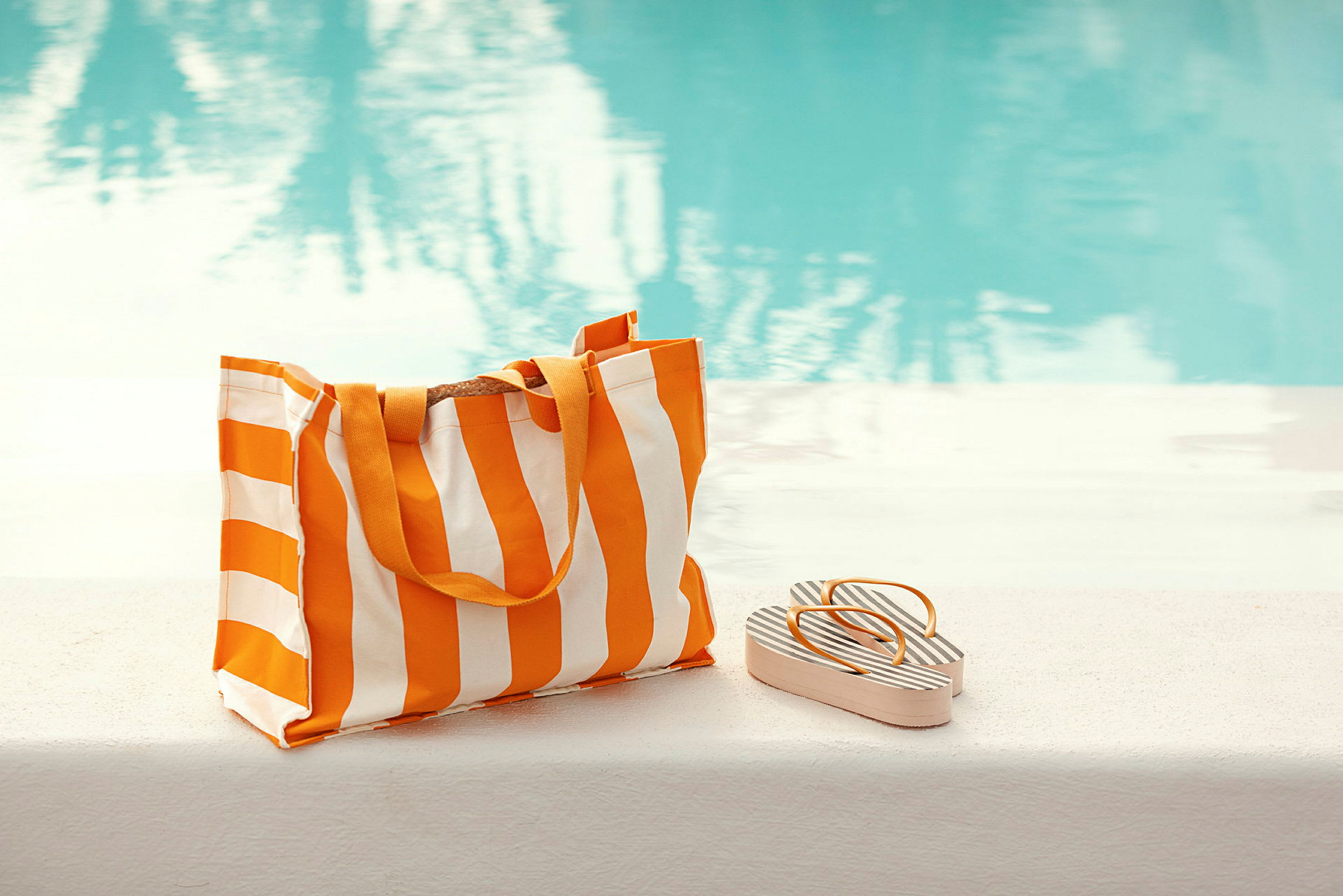 En stripete strandveske og sandaler ligger ved siden av et basseng. (AI-generert tekst)