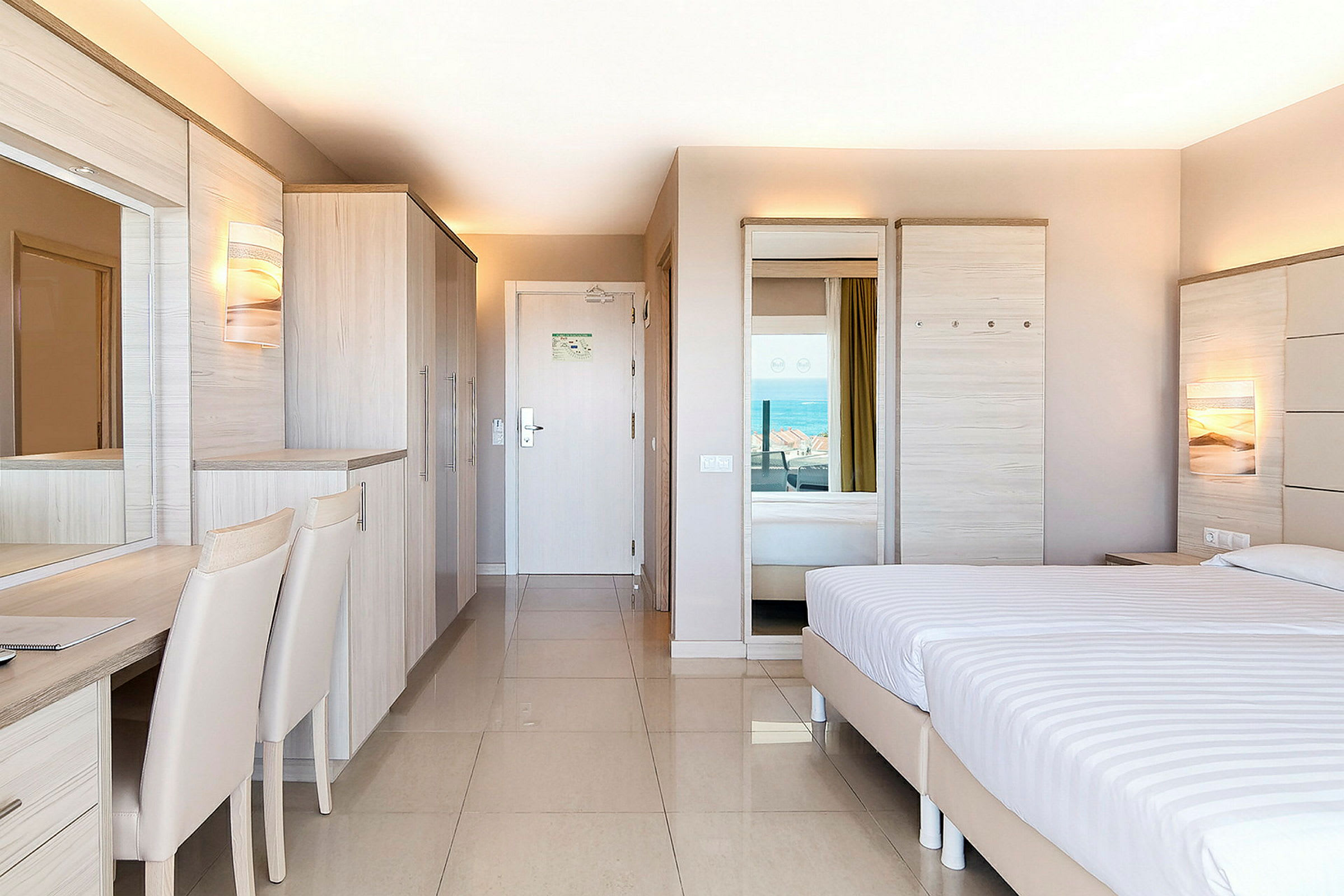 Et hotellrom med moderne innredning, en dobbeltseng, garderobe og skrivebord, med utsikt mot havet. (AI-generert tekst)