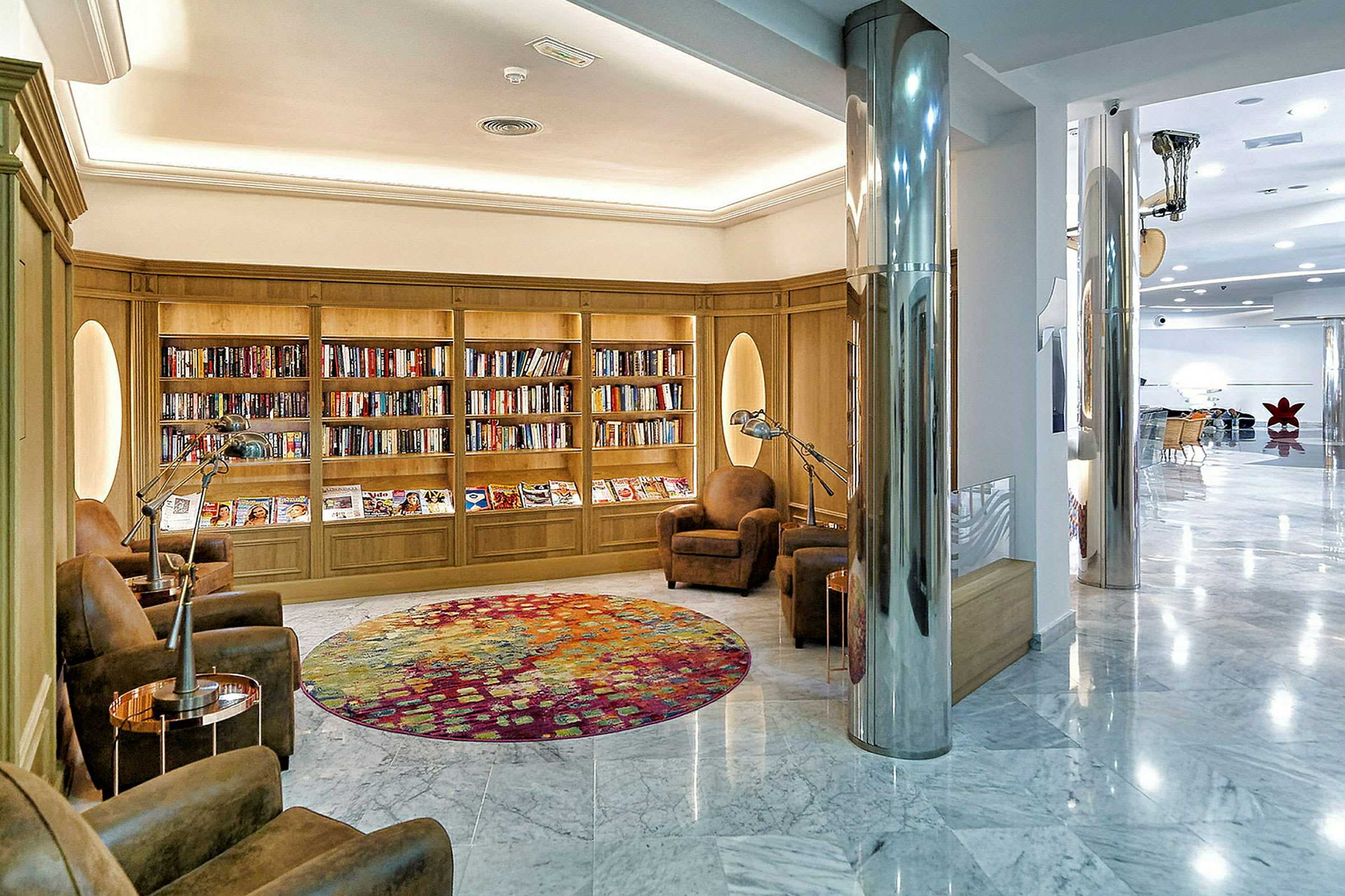 Et elegant hotellbibliotek med bokhyller, komfortable lenestoler og et fargerikt rundt gulvteppe. (AI-generert tekst)