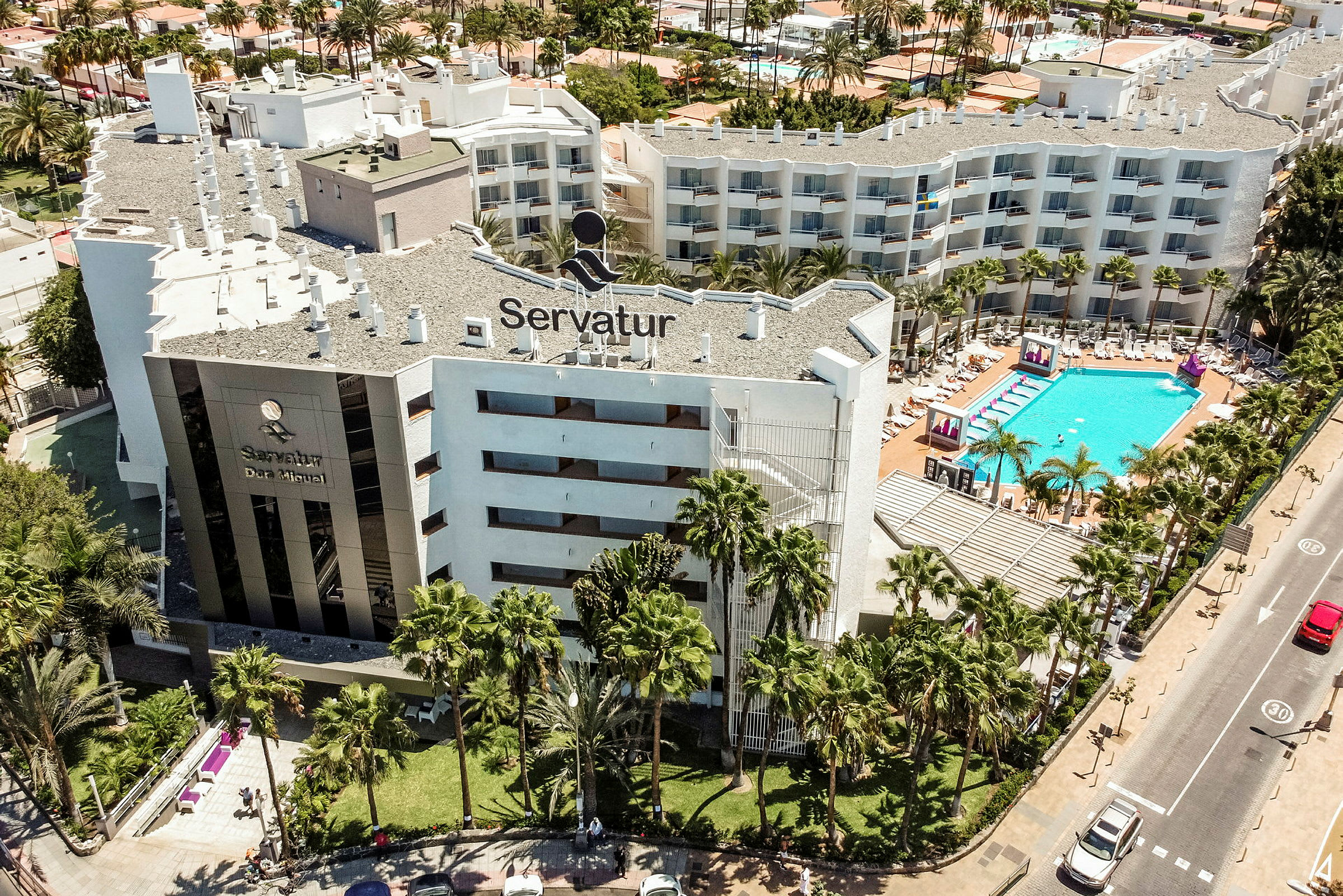 Luftfoto av hotellbygning med bassengområde og omkringliggende grøntområder. (AI-generert tekst)