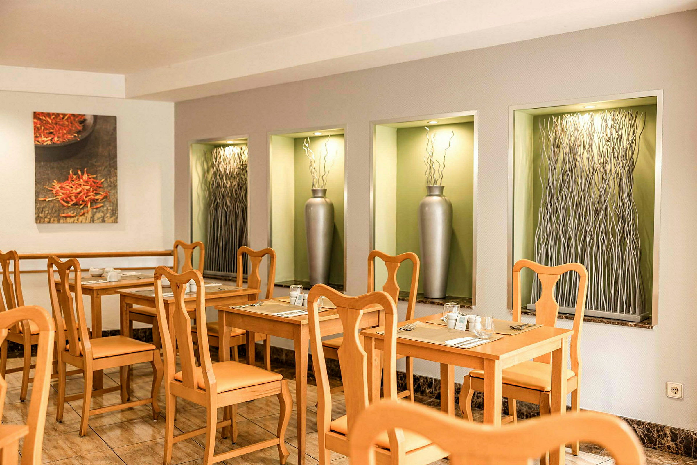 Et restaurantområde med trebord og stoler, samt dekorative nisjer i veggen med vaser og kunstverk. (AI-generert tekst)