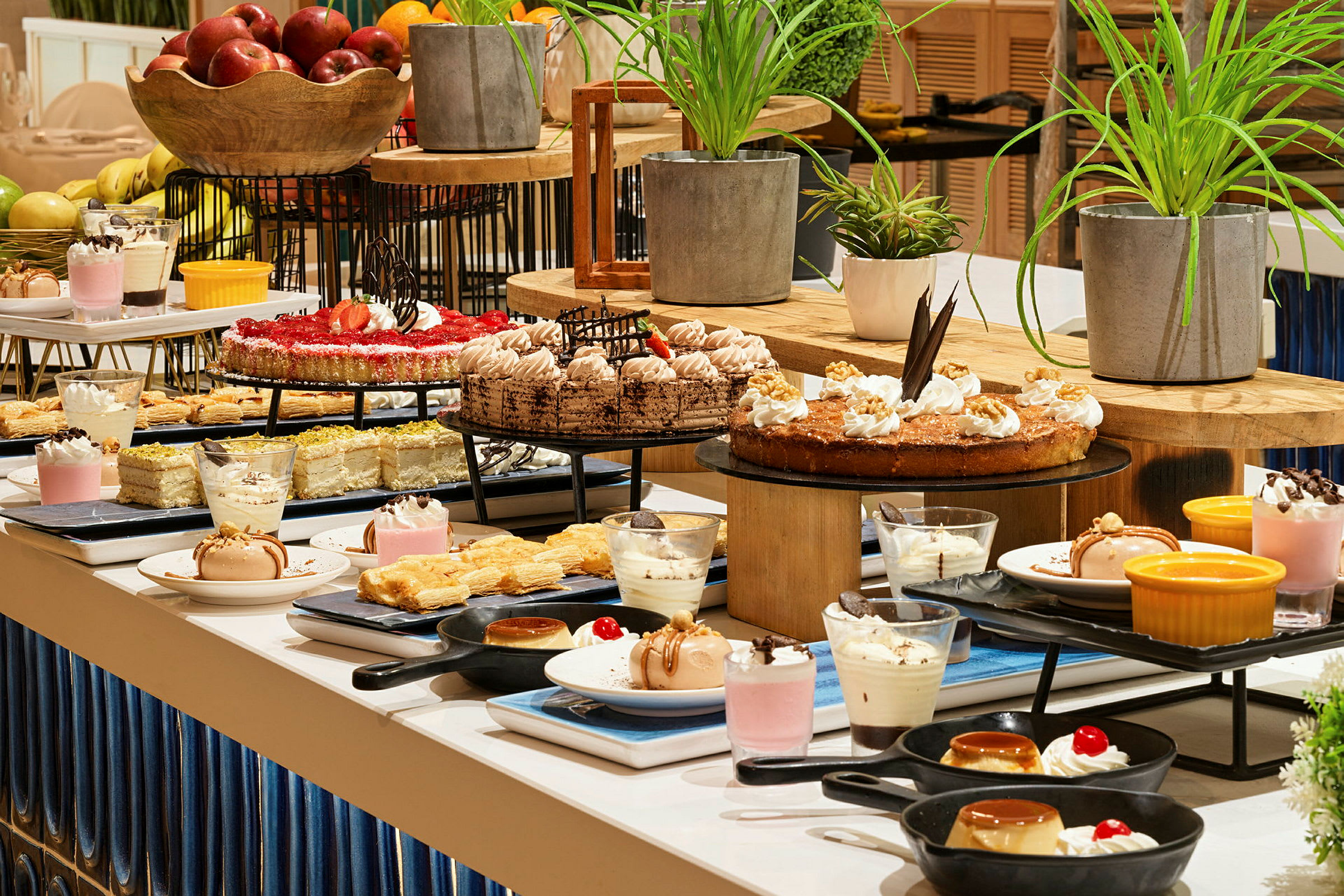 En buffé med ulike desserter, inkludert kaker, bakverk, mousse og frukt. Planter er plassert som dekorasjon. (AI-generert tekst)