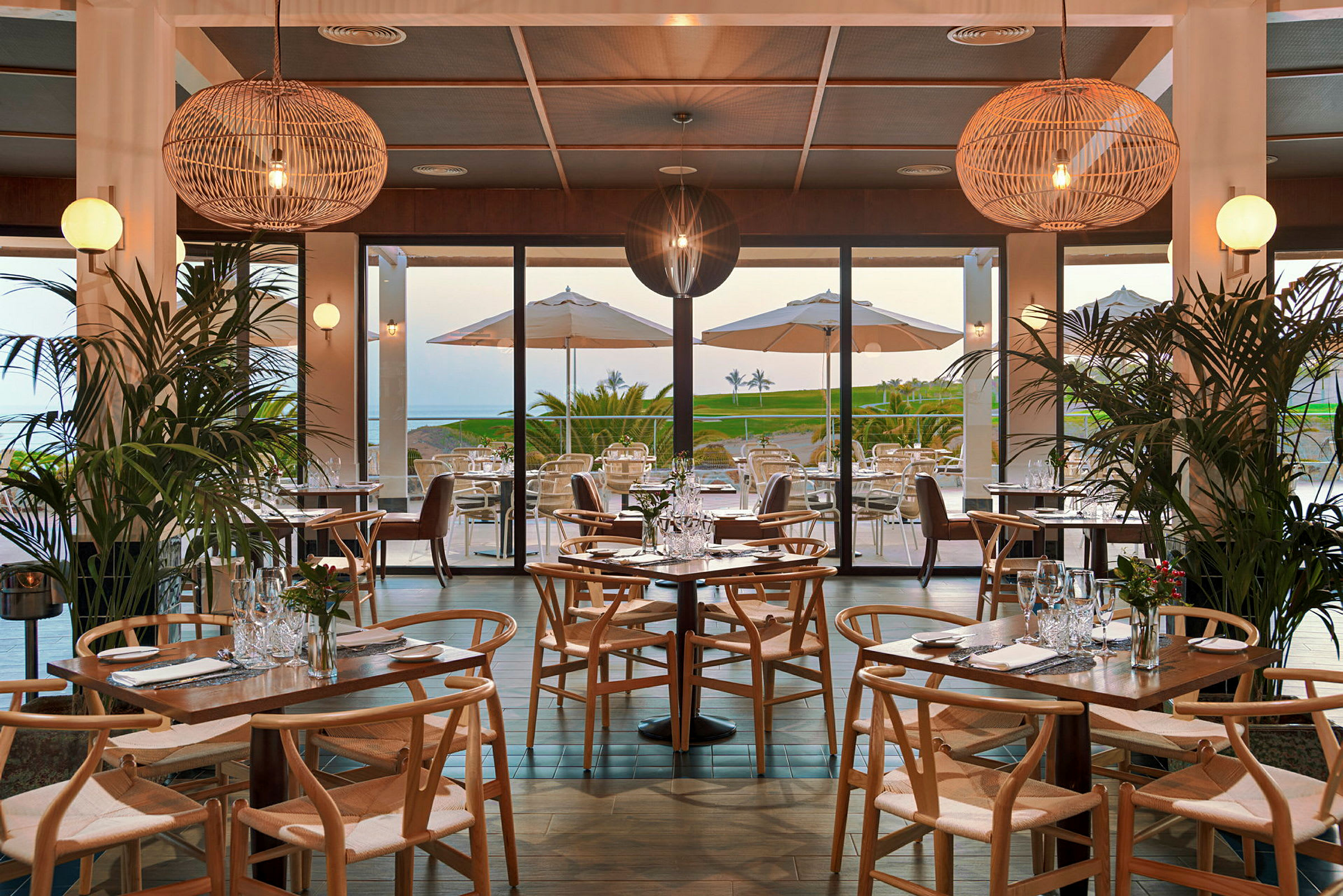 En restaurant med trebord og stoler, dekorert med planter og store hengende lamper. I bakgrunnen kan man se en terrasse med parasoller og utsikt mot et grønt område og havet. (AI-generert tekst)