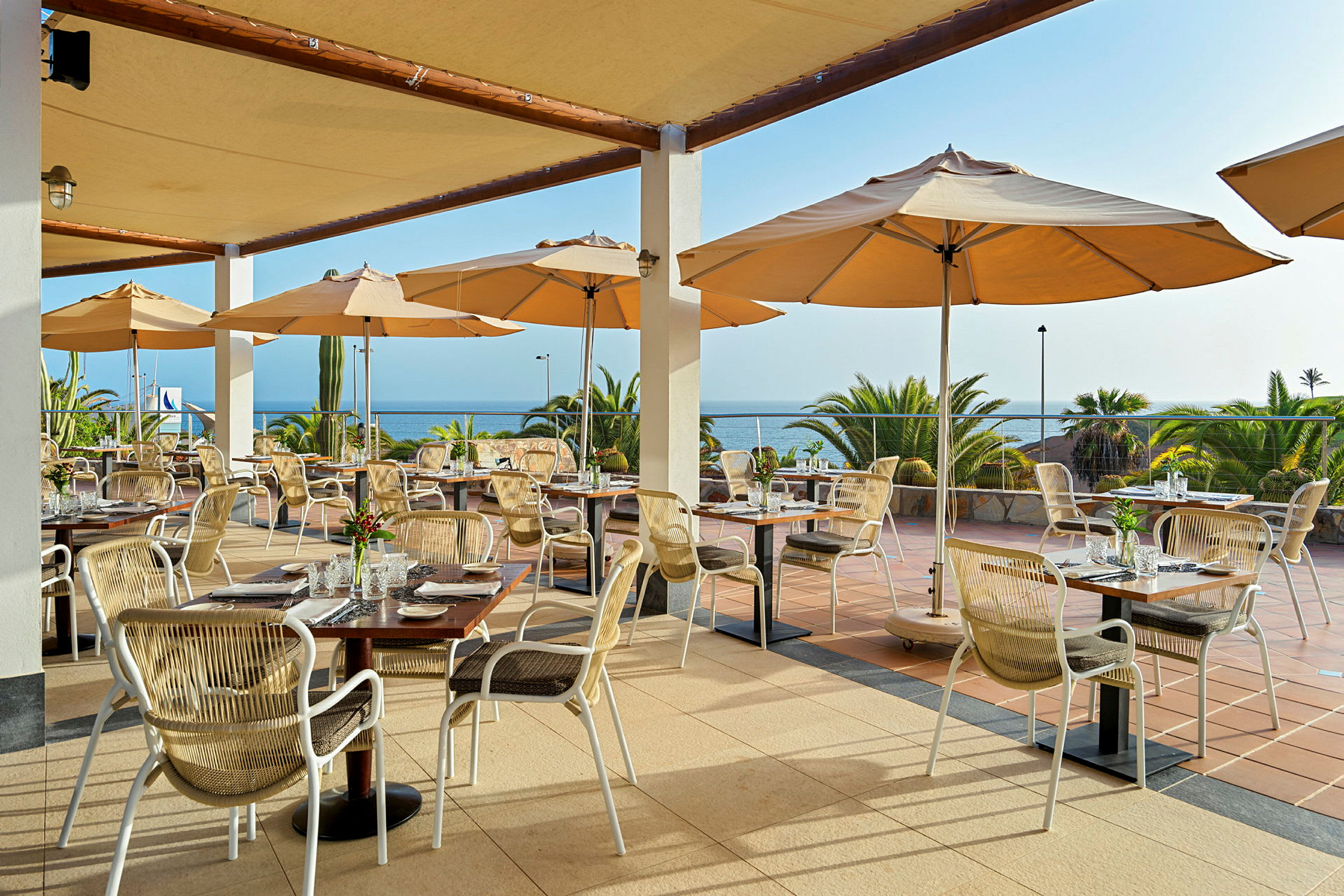 Restaurant med uteservering, bord og stoler under parasoller med utsikt over havet. (AI-generert tekst)