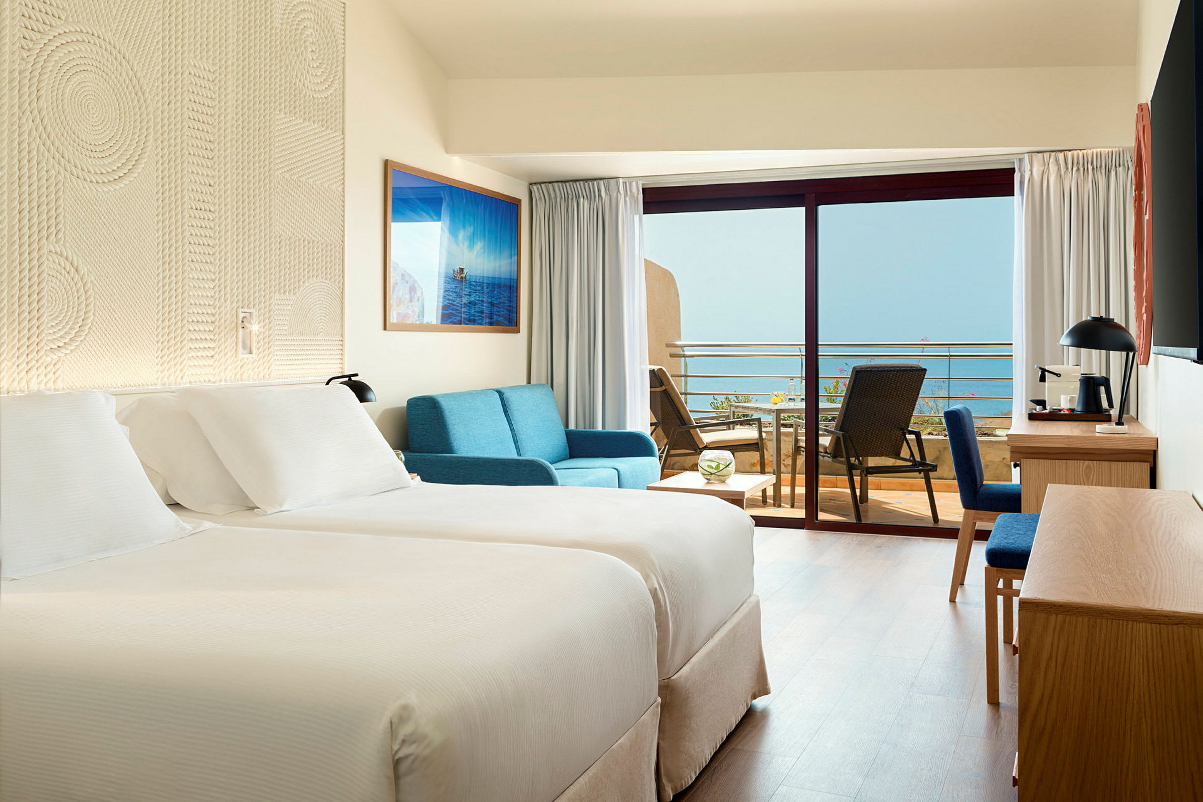 Et hotellrom med to enkeltsenger, en sofa, et skrivebord og en balkong med utsikt over havet. (AI-generert tekst)