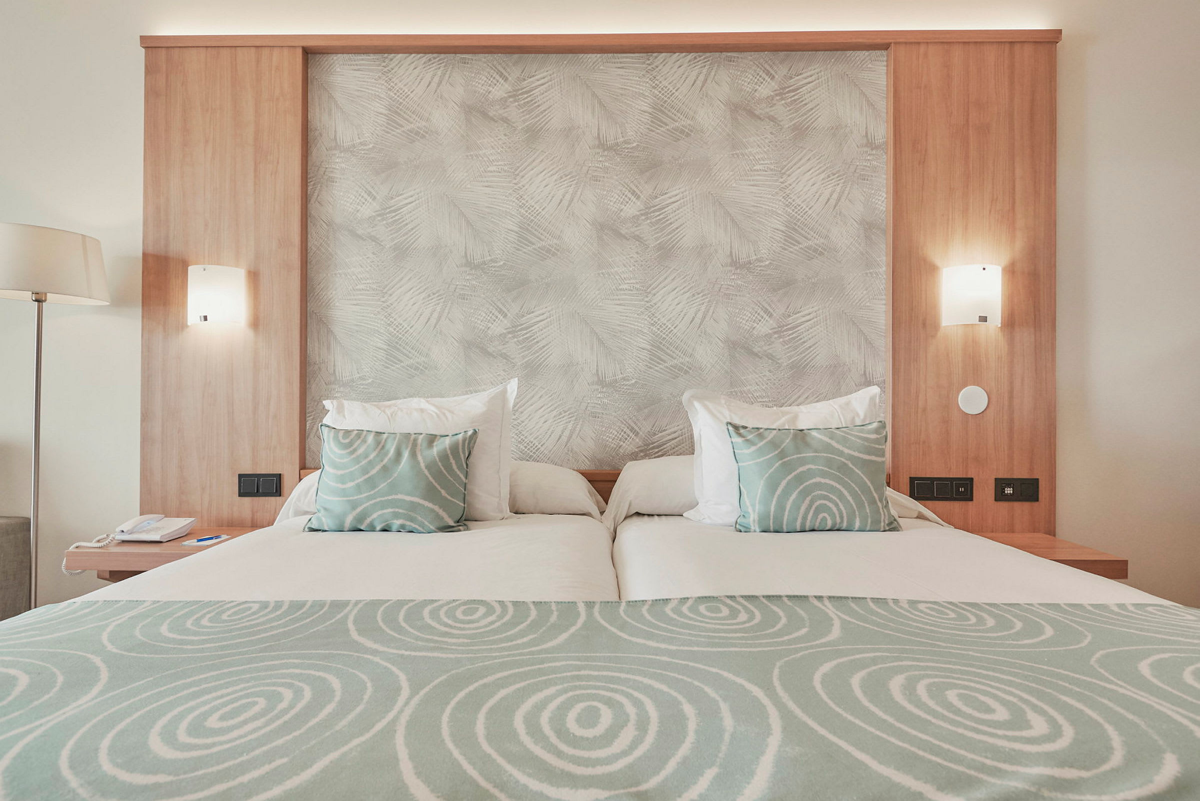 Hotellseng med lys trepanel, grønne puter med sirkelmønster og matchende sengetøy. (AI-generert tekst)