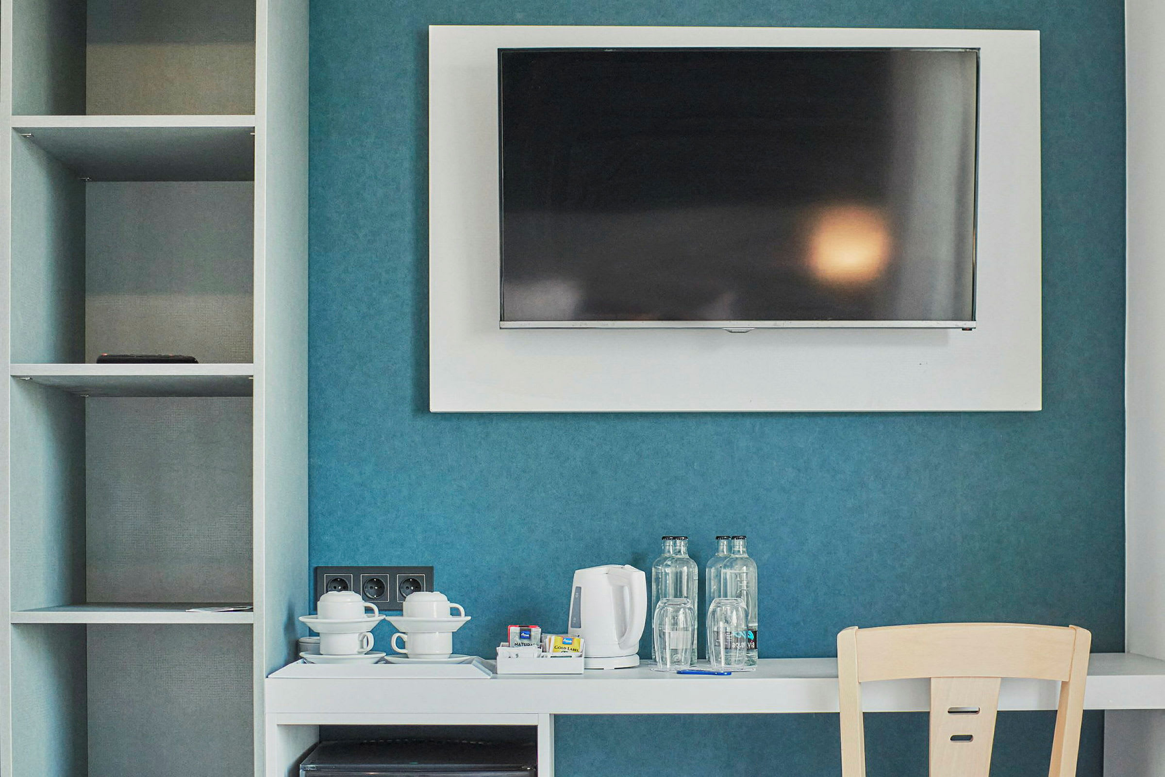 Et hotellrom med en veggmontert TV, en hvit møbel med vannkoker, kopper og flasker, samt et åpent skap på siden. (AI-generert tekst)