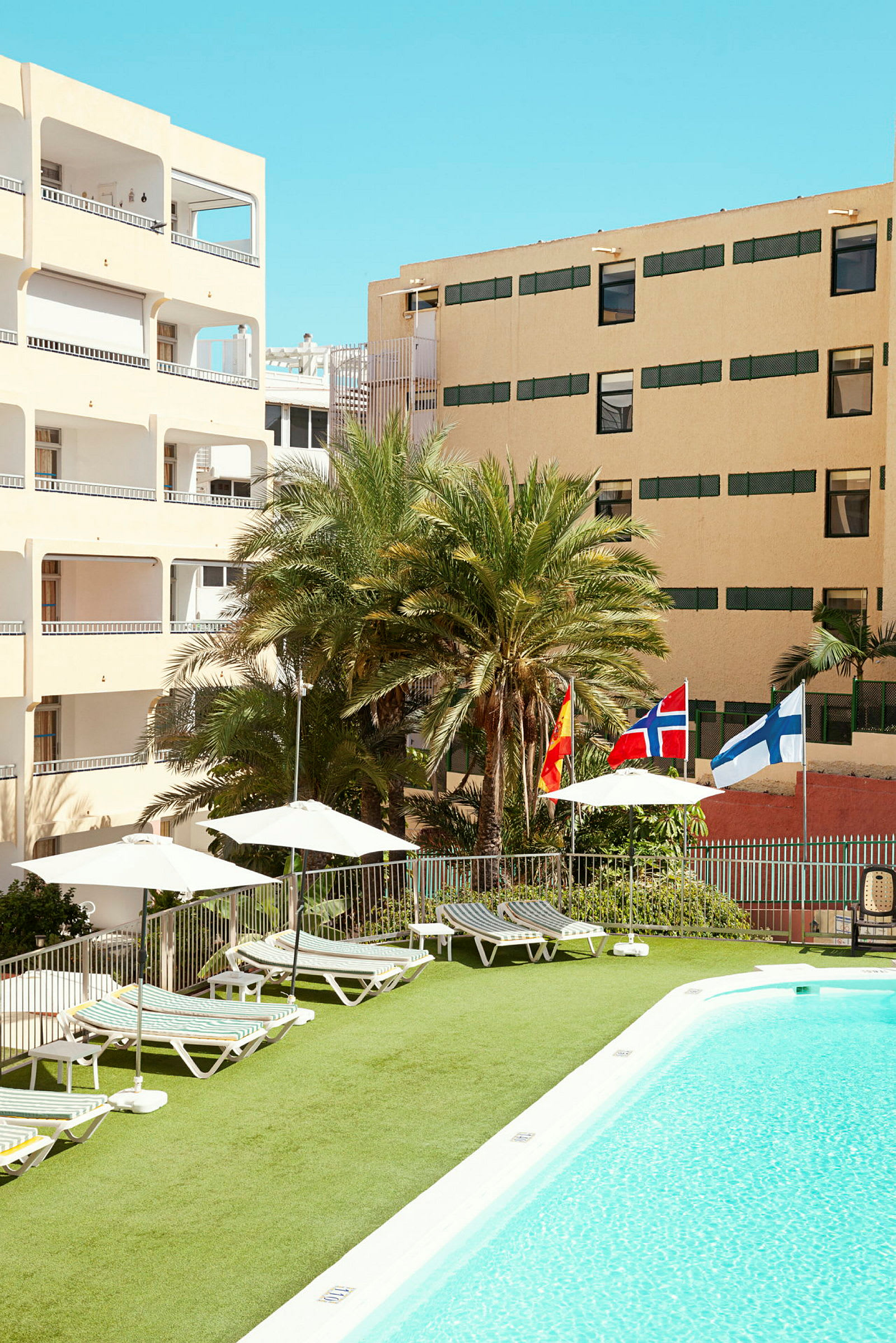Bilde av et hotellbasseng med solsenger, parasoller og flagg fra Spania, Norge og Finland. Bakgrunnen viser hotellbygninger og palmer. (AI-generert tekst)