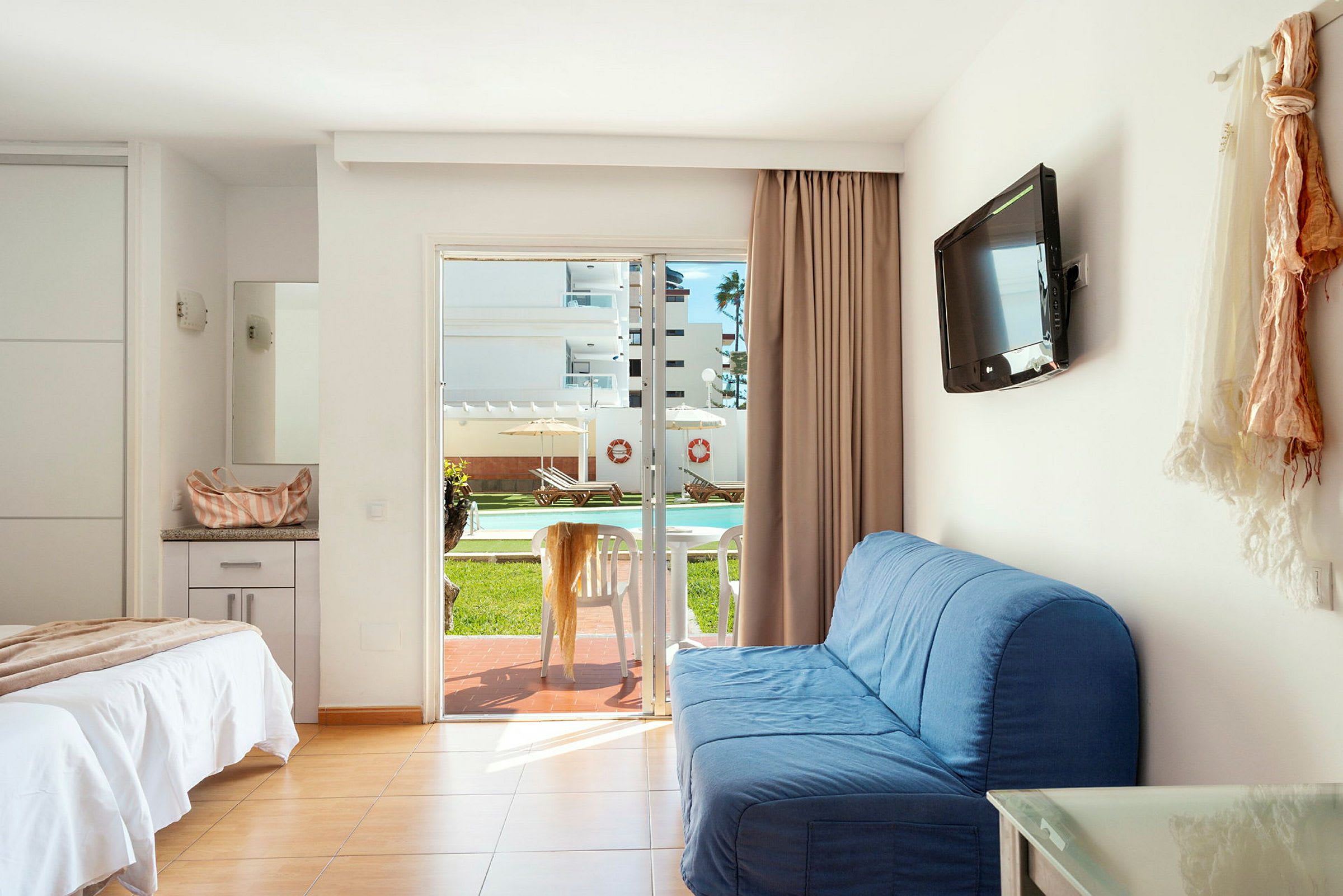 Et hotellrom med en oppredd seng, blå sofa, veggmontert TV og en glassdør som fører ut til en terrasse med bord og stoler. Utsikten viser et utendørsbasseng og solsenger. (AI-generert tekst)