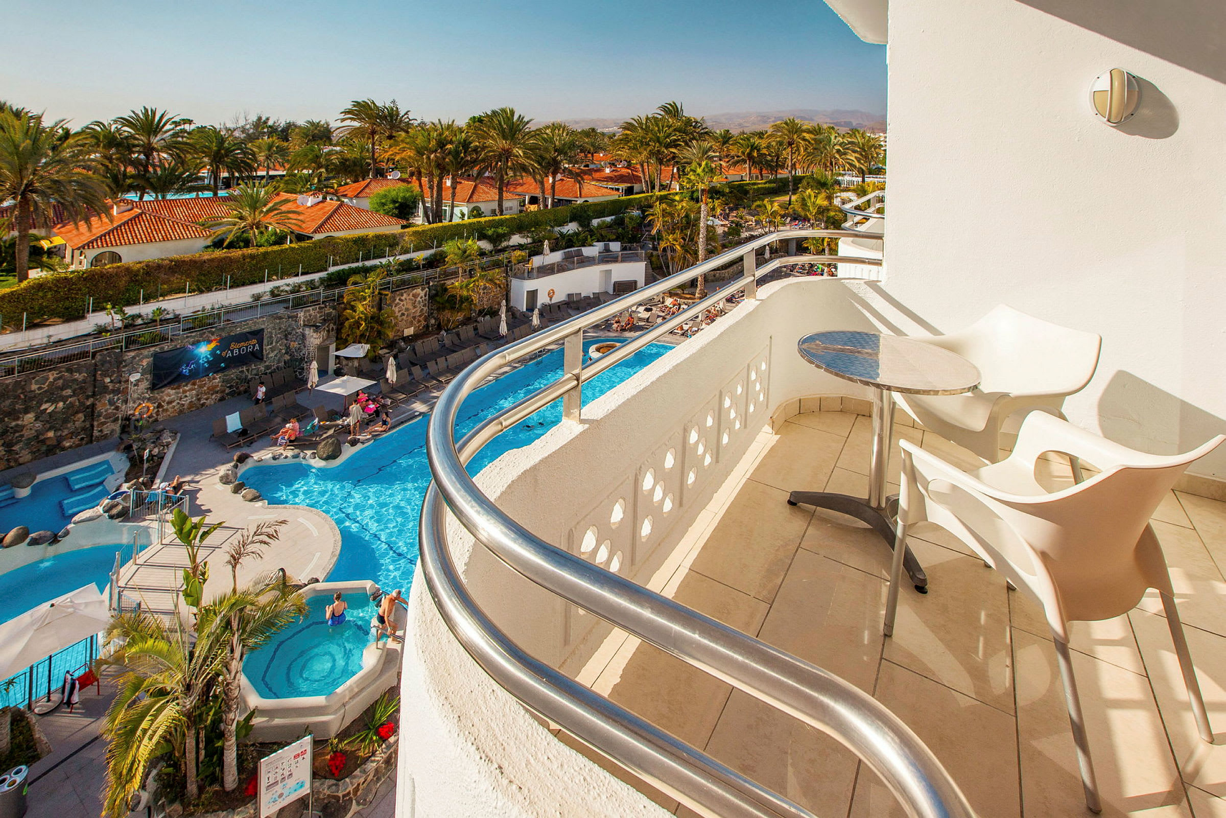 Utsikt fra en balkong med bord og stoler mot et basseng og palmer. (AI-generert tekst)