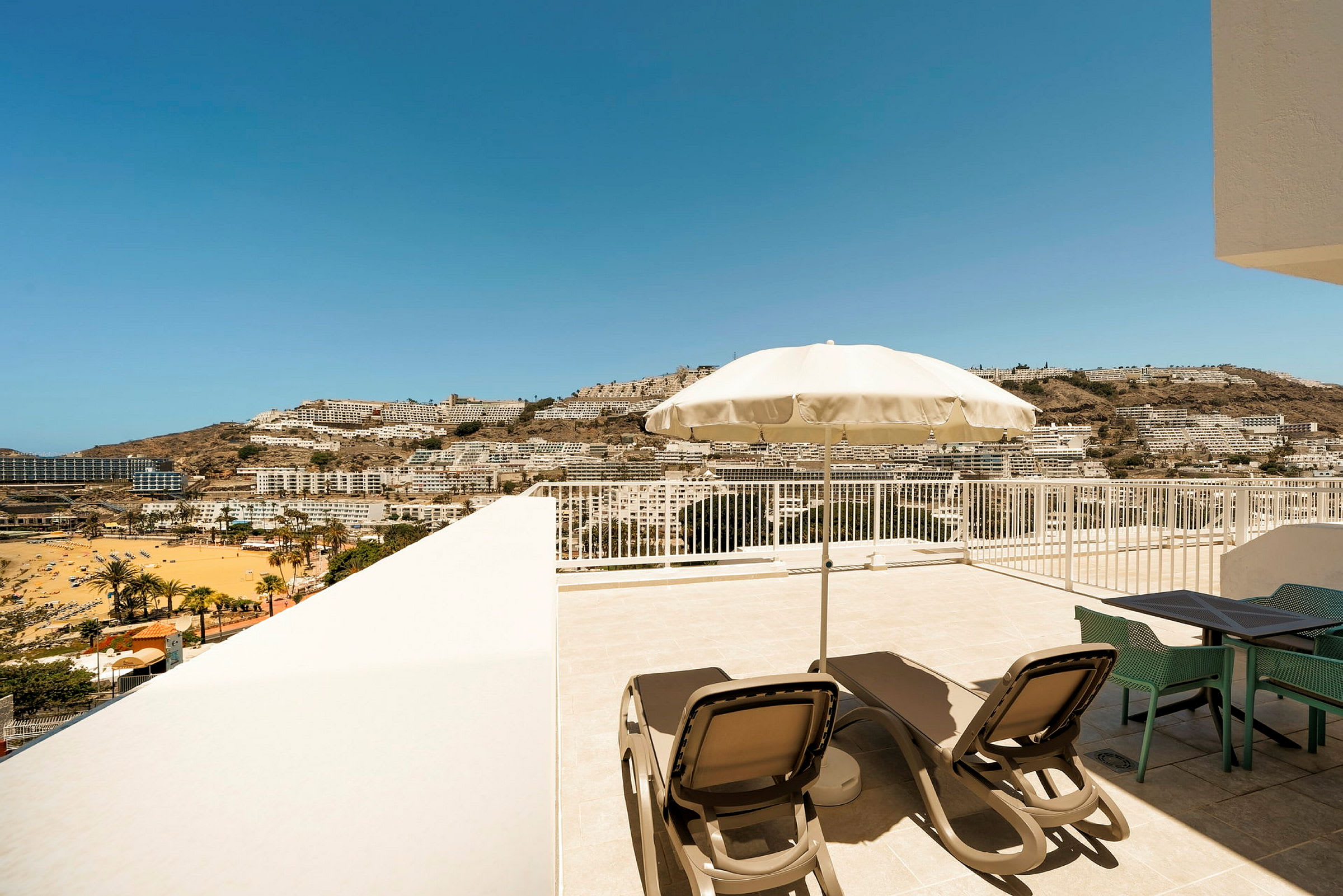 Terrasse med solsenger og parasoll, utsikt over en strand og by mot fjell i det fjerne. (AI-generert tekst)