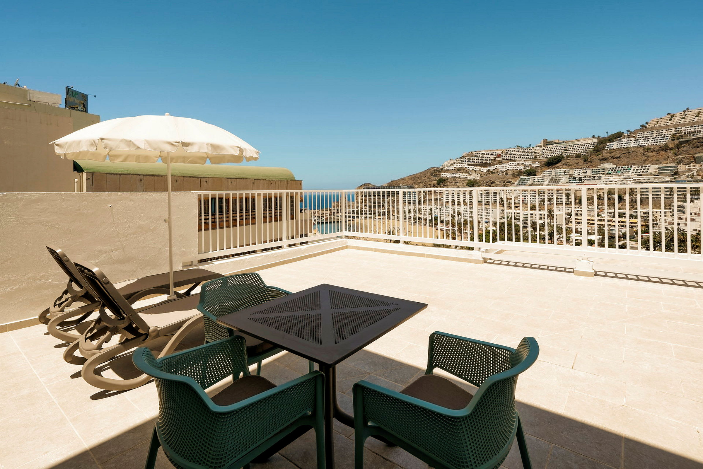Utsikt fra en terrasse med bord, stoler og solsenger, mot havet og bygninger på en fjellskråning. (AI-generert tekst)