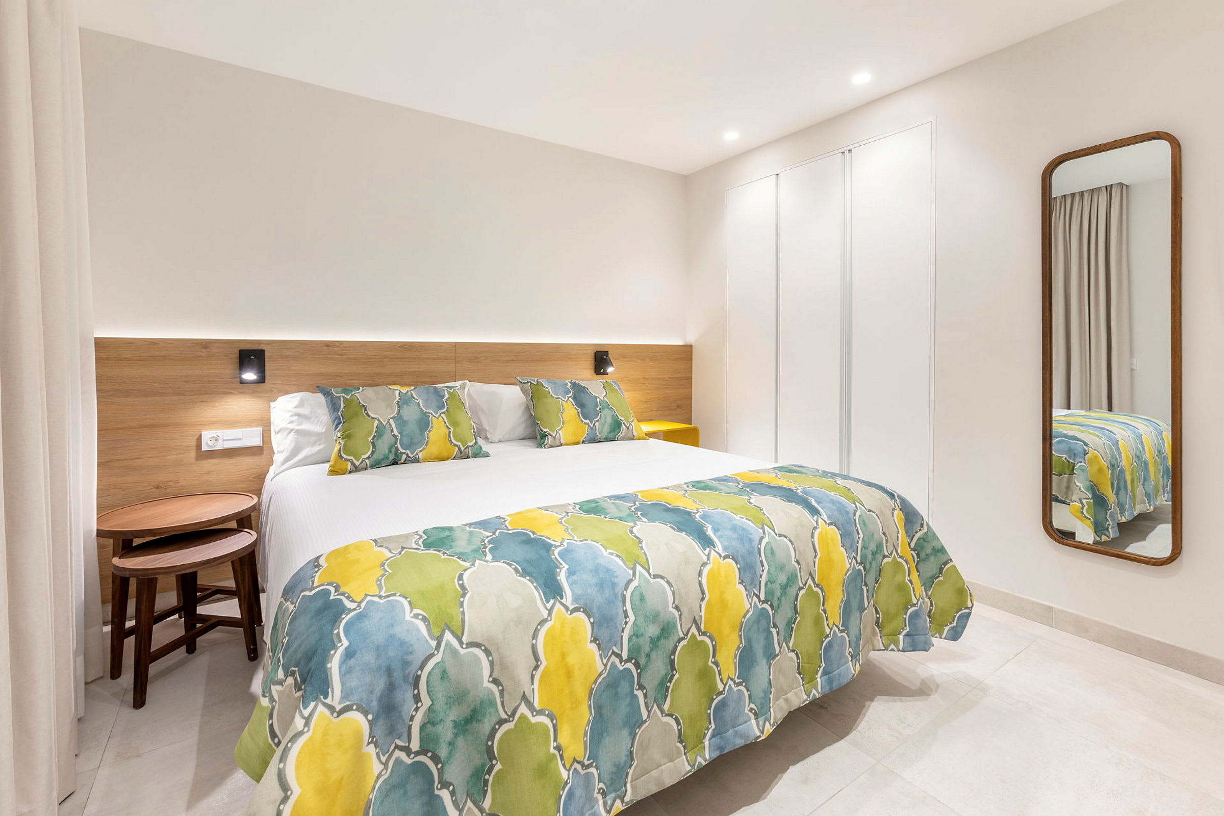 Et moderne hotellrom med en seng dekket med et fargerikt sengeteppe og puter, samt et speil på veggen. (AI-generert tekst)