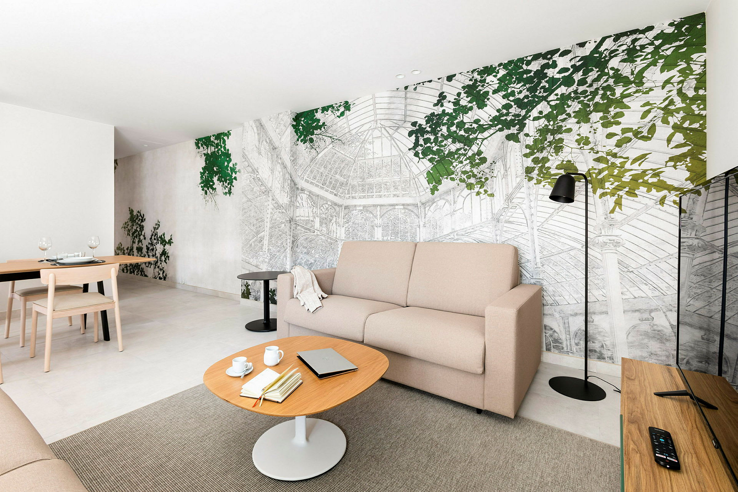 Et oppholdsrom med en beige sofa, et trebord og en veggdekorasjon som forestiller et drivhus med grønne blader. (AI-generert tekst)