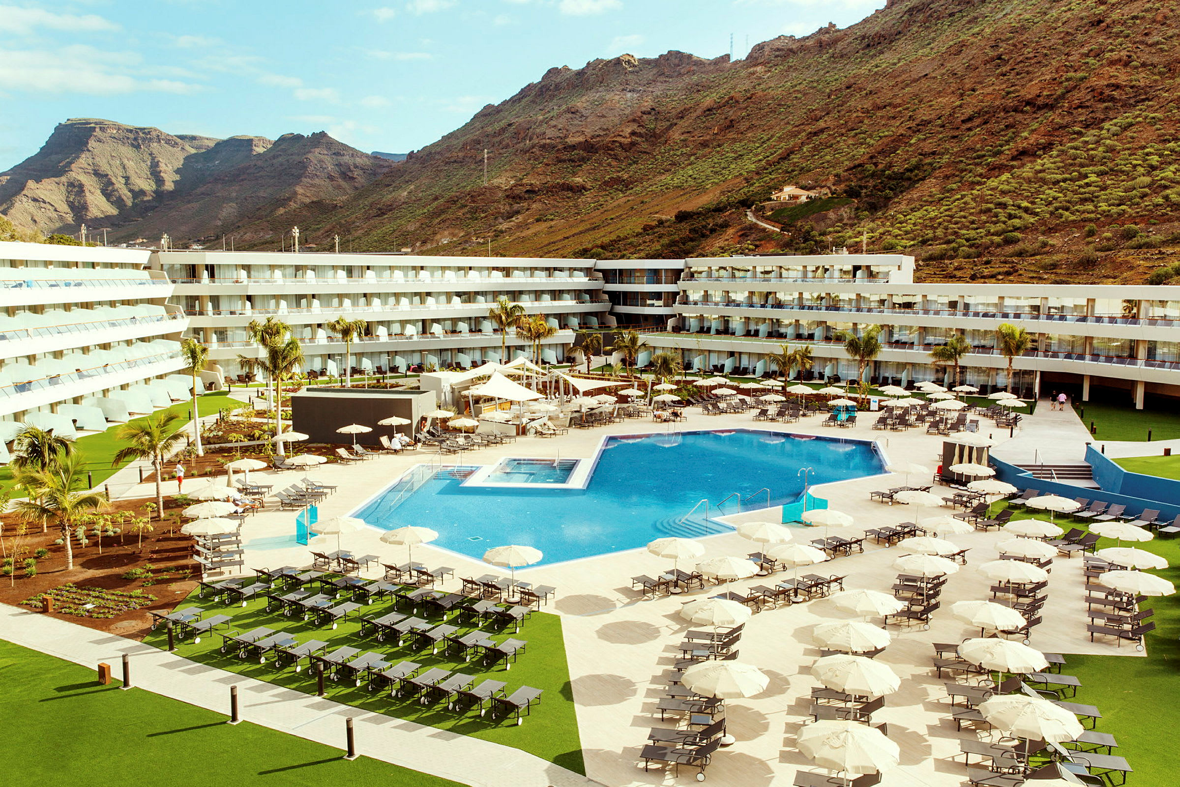 Et hotellområde med et stort utendørsbasseng, solsenger og parasoller, omgitt av bygninger og fjell i bakgrunnen. (AI-generert tekst)