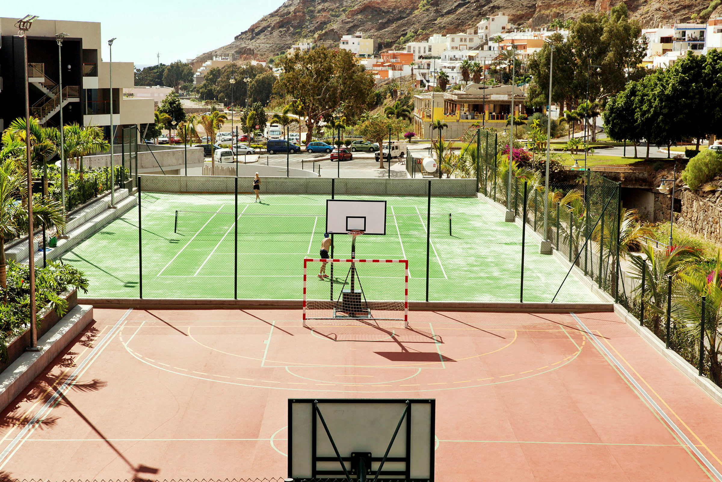 Et bilde av en fargerik idrettsanlegg med basketballmål og tennisbaner, omgitt av grøntområder, bygninger og åser. (AI-generert tekst)
