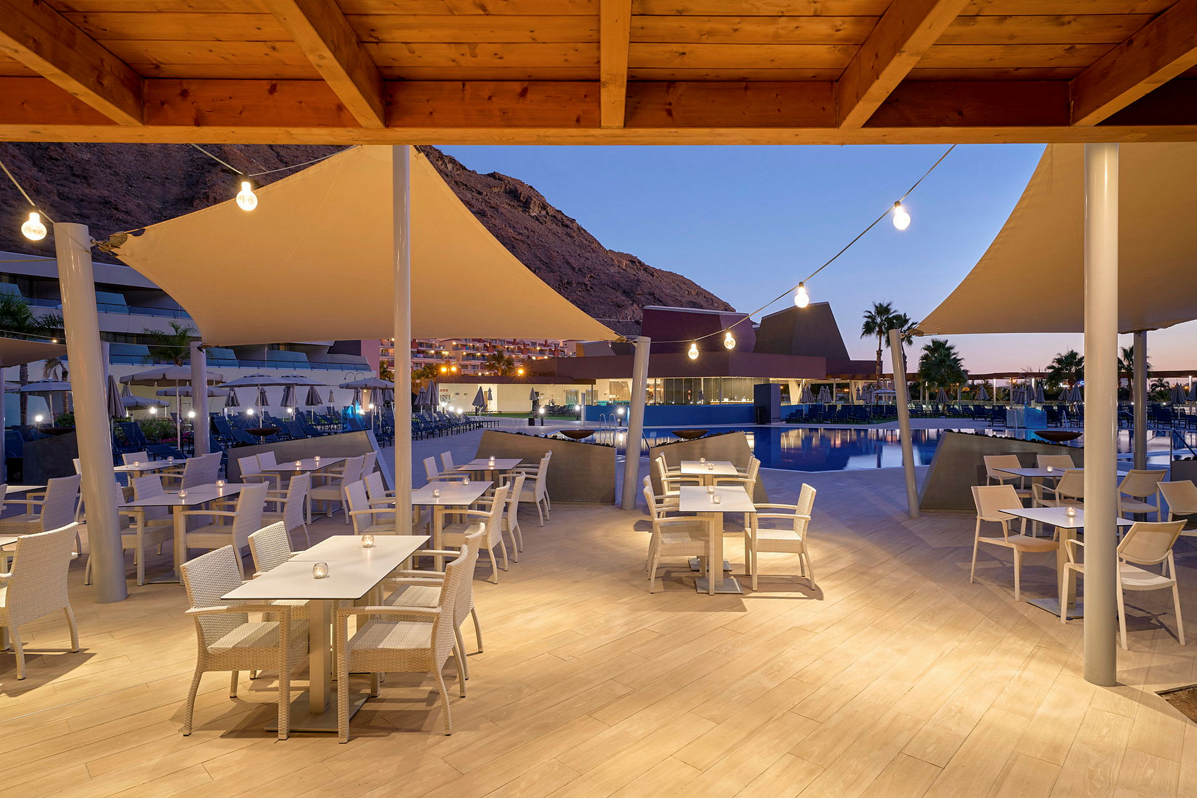 Utendørs restaurant med hvite stoler og bord ved et basseng, kveldsbelysning og fjell i bakgrunnen. (AI-generert tekst)