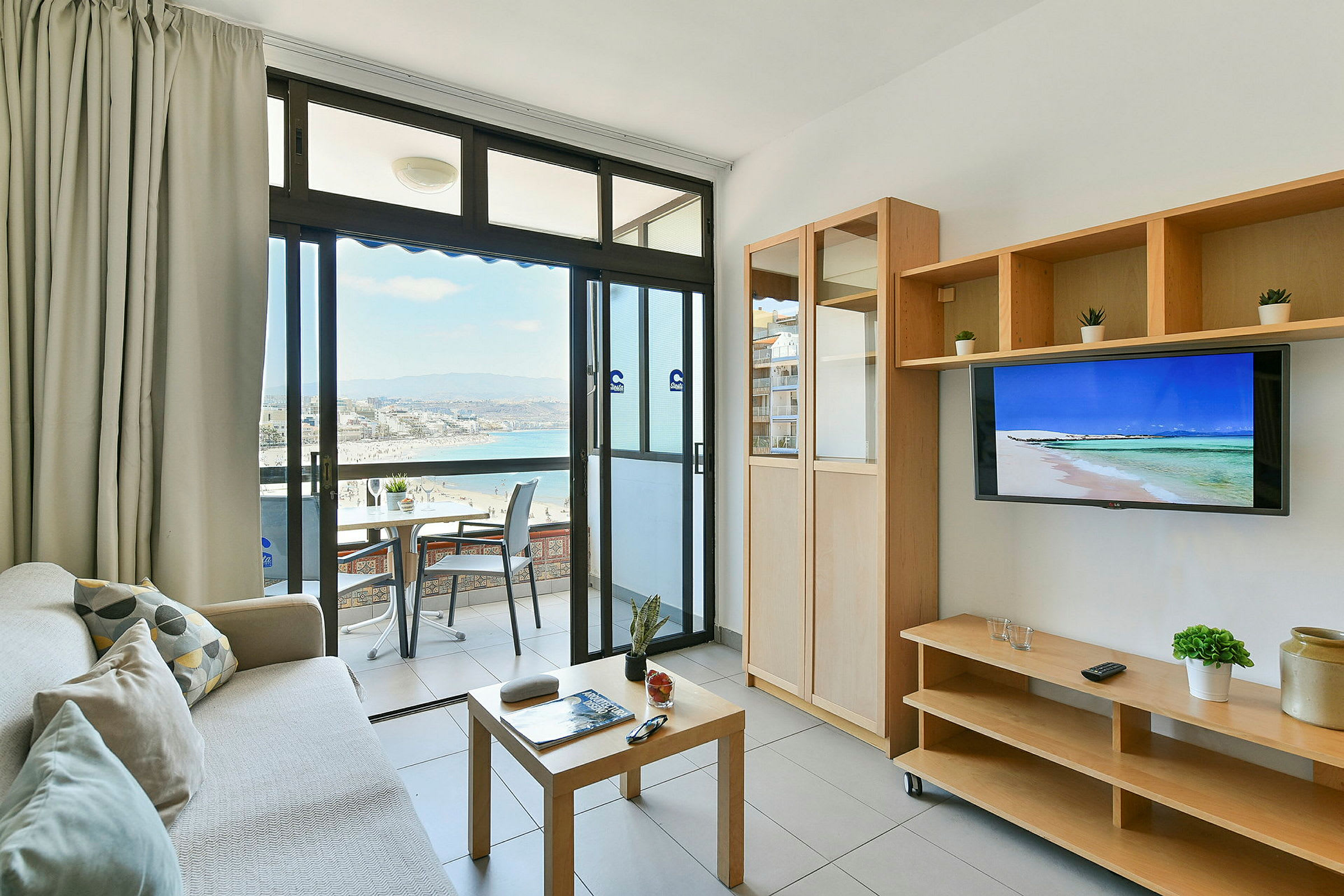 En stue med utgang til en balkong som har utsikt over en strand og sjøen. Rommet har møbler som sofa, salongbord og en TV. (AI-generert tekst)