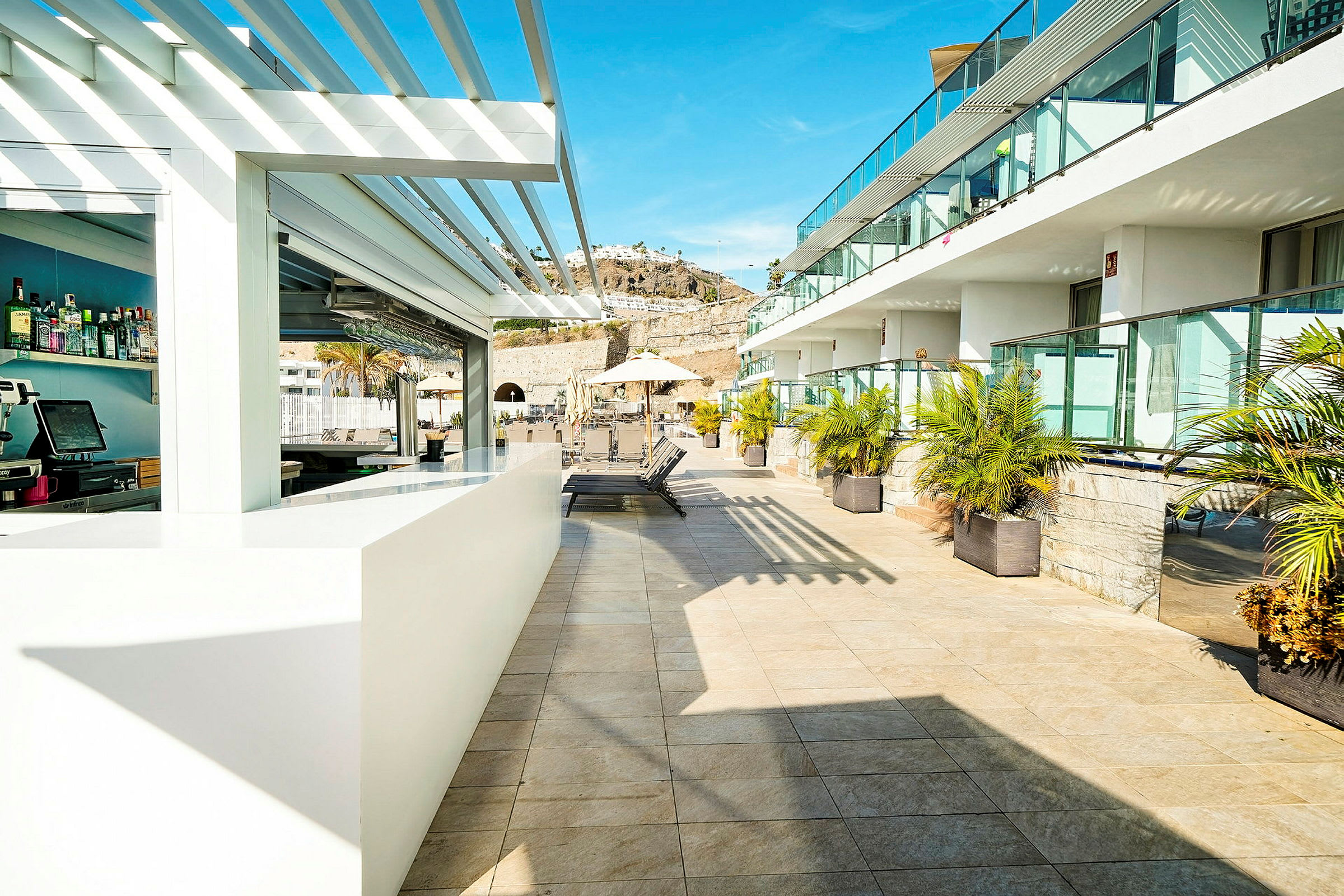 Bilde av en solrik hotellterrasse med et barområde og solsenger, omgitt av palmer og glassrekkverk. (AI-generert tekst)