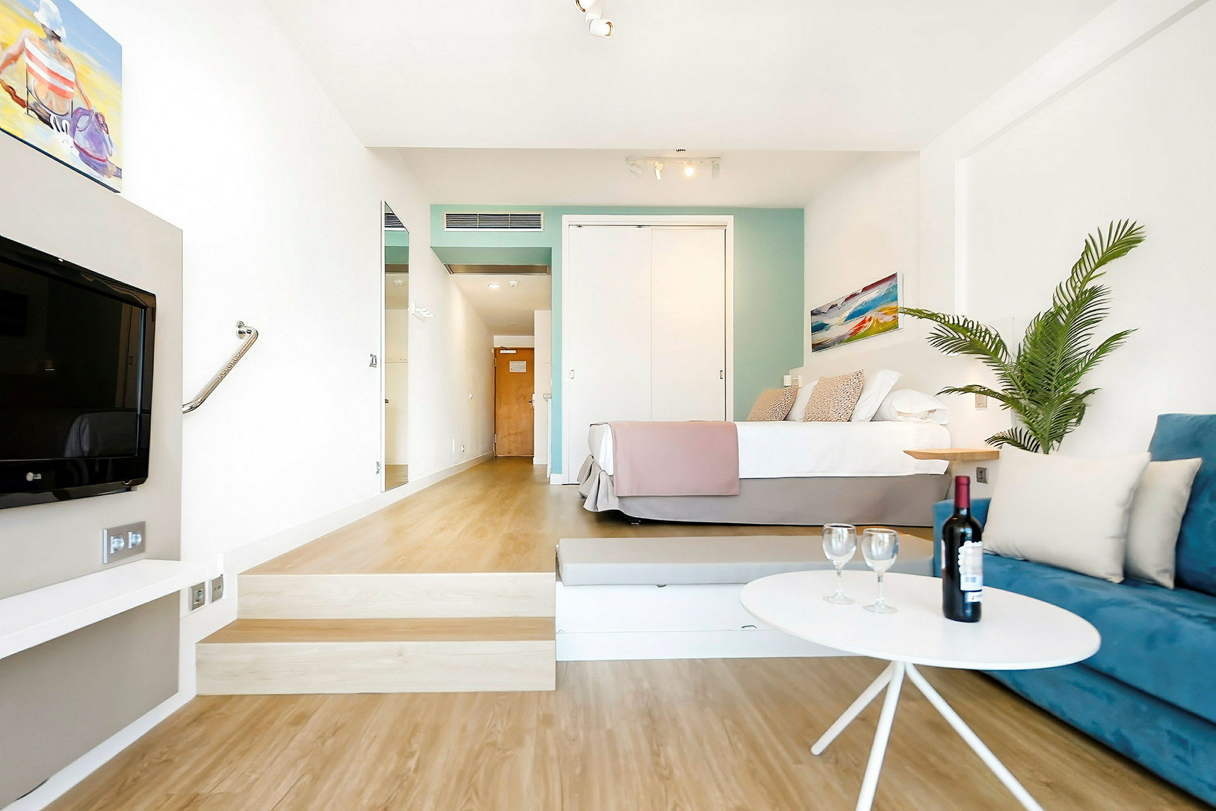 Et hotellrom med en seng, en blå sofa, et bord med en vinflaske og glass, og en plante. (AI-generert tekst)