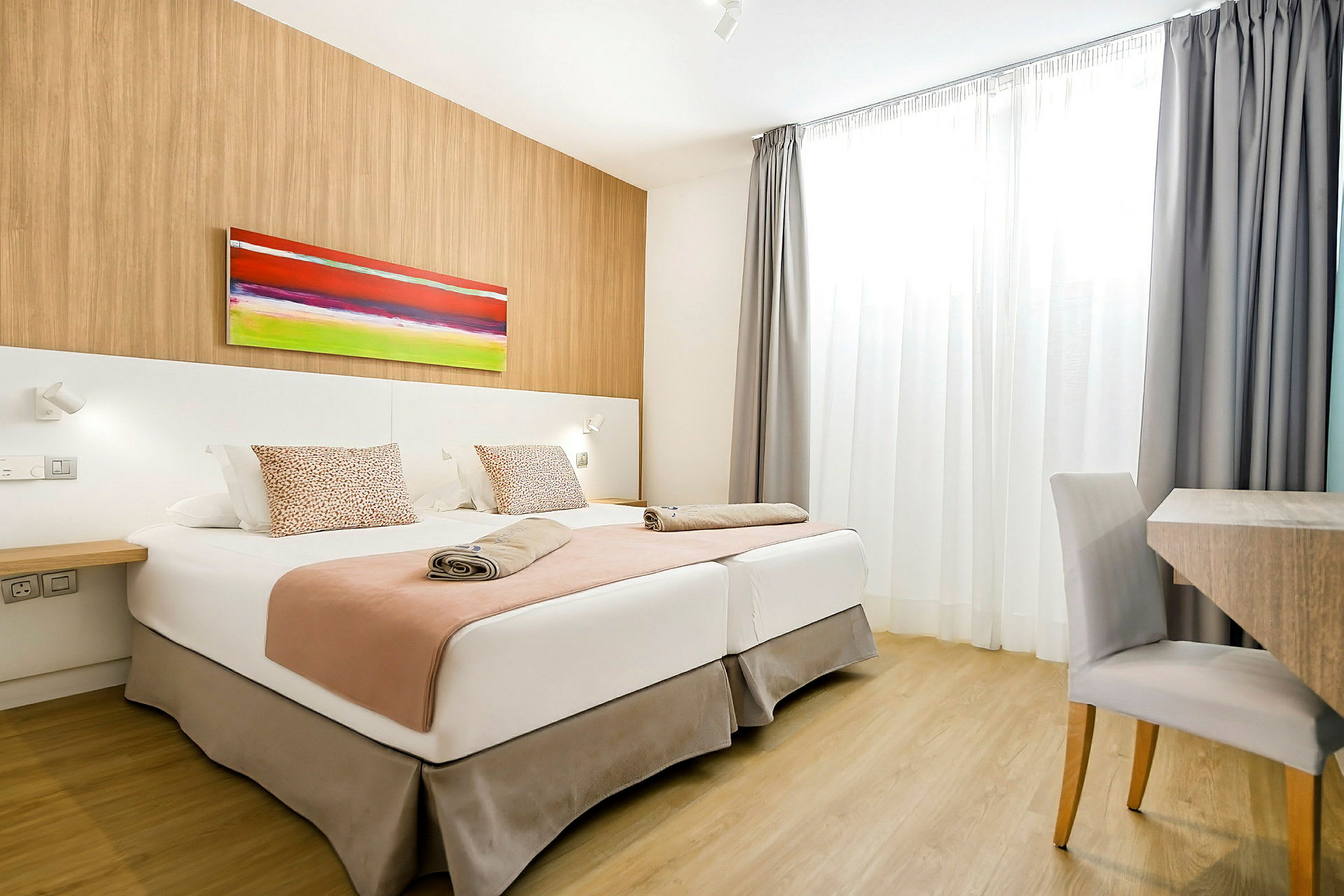 Et hotellrom med to senger, et fargerikt maleri på veggen og et skrivebord med stol. (AI-generert tekst)