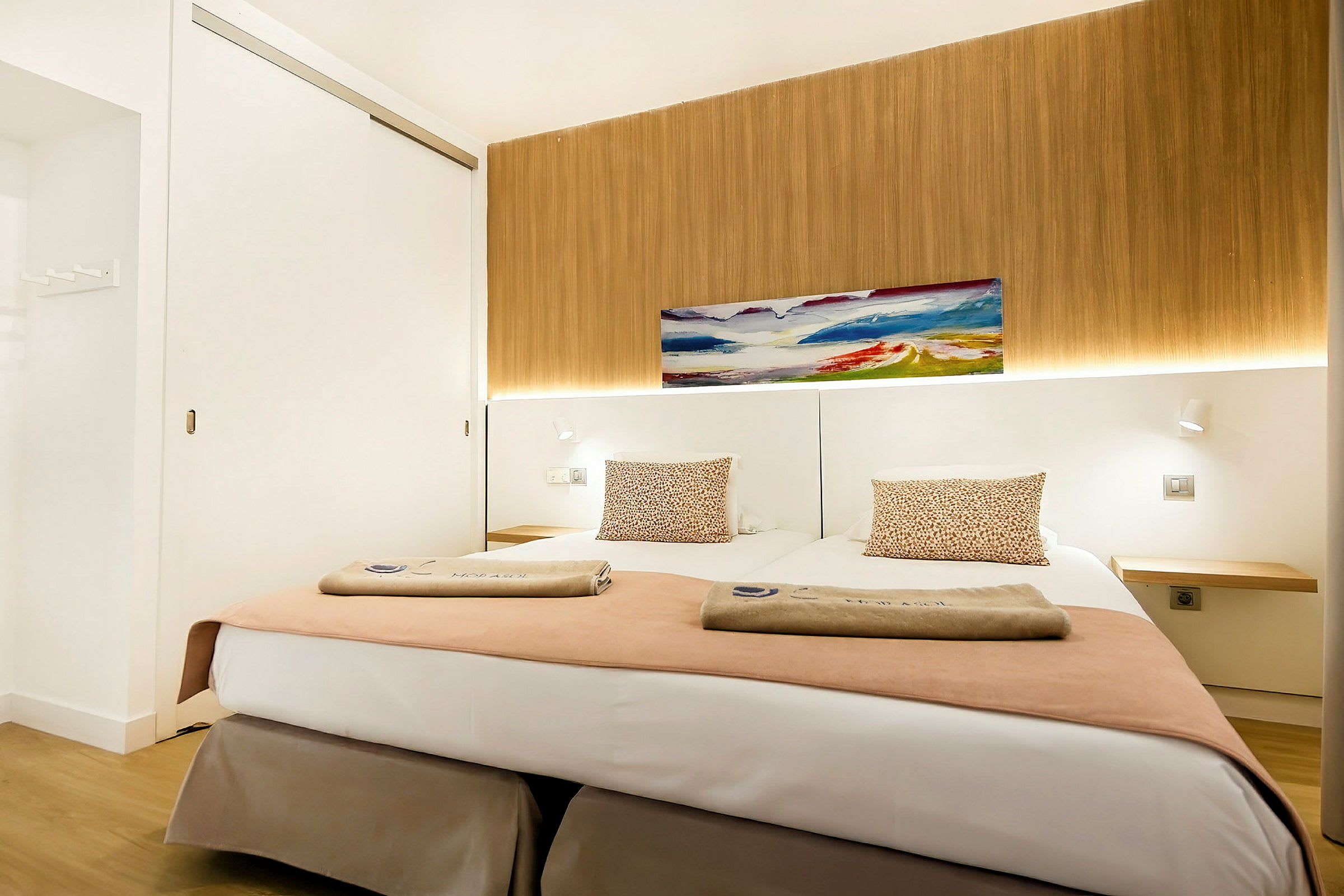 Et moderne hotellrom med to enkeltsenger, dekorert med puter og håndklær, et maleri på veggen og et trebakgrunnspanel. (AI-generert tekst)
