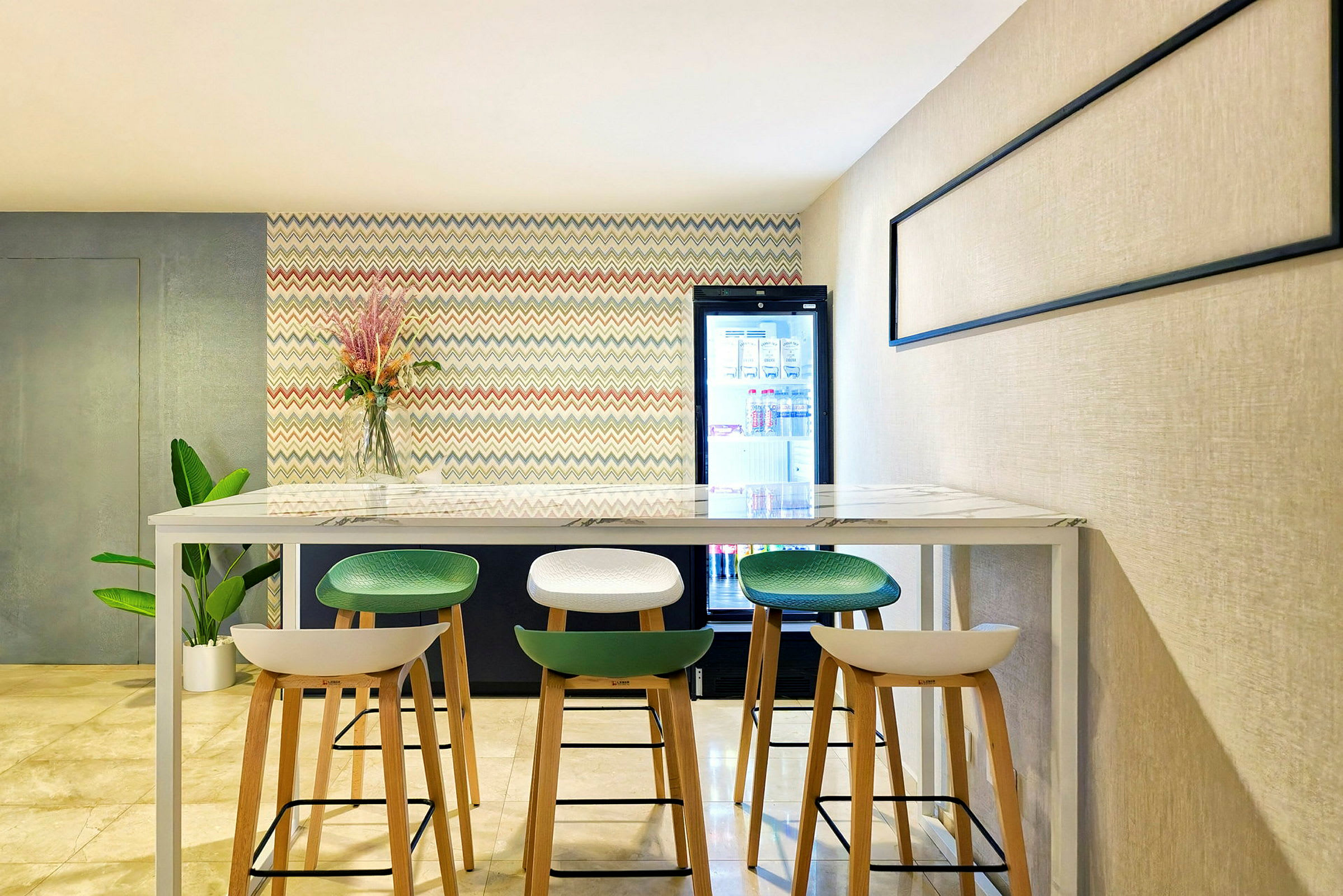 Et moderne rom med høye stoler plassert rundt et langbord. På veggen bak er et fargerikt mønster og en glassvase med blomster. Til høyre er et kjøleskap. (AI-generert tekst)