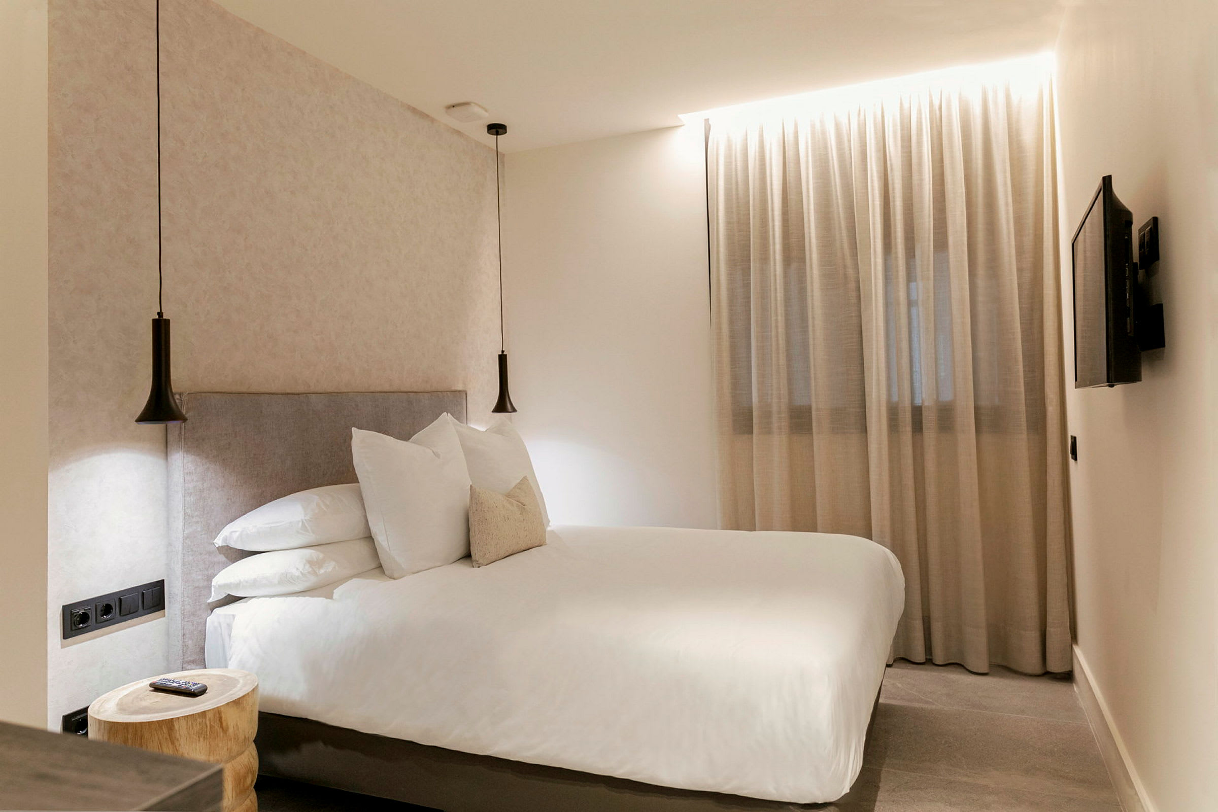 Et hotellrom med en dobbeltseng, to hengende lamper, et vindu med gjennomsiktige gardiner og en veggmontert TV. (AI-generert tekst)