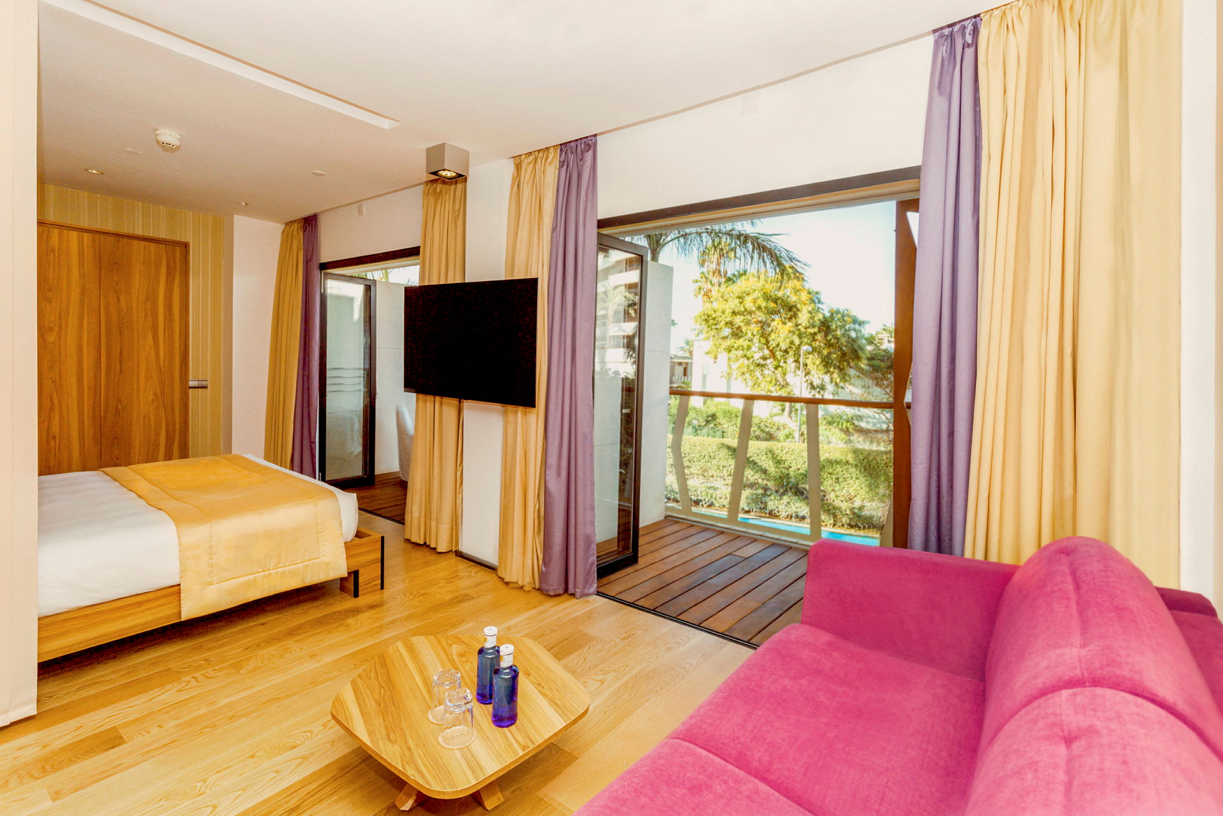 Hotellrom med seng, rosa sofa, TV og balkong. (AI-generert tekst)