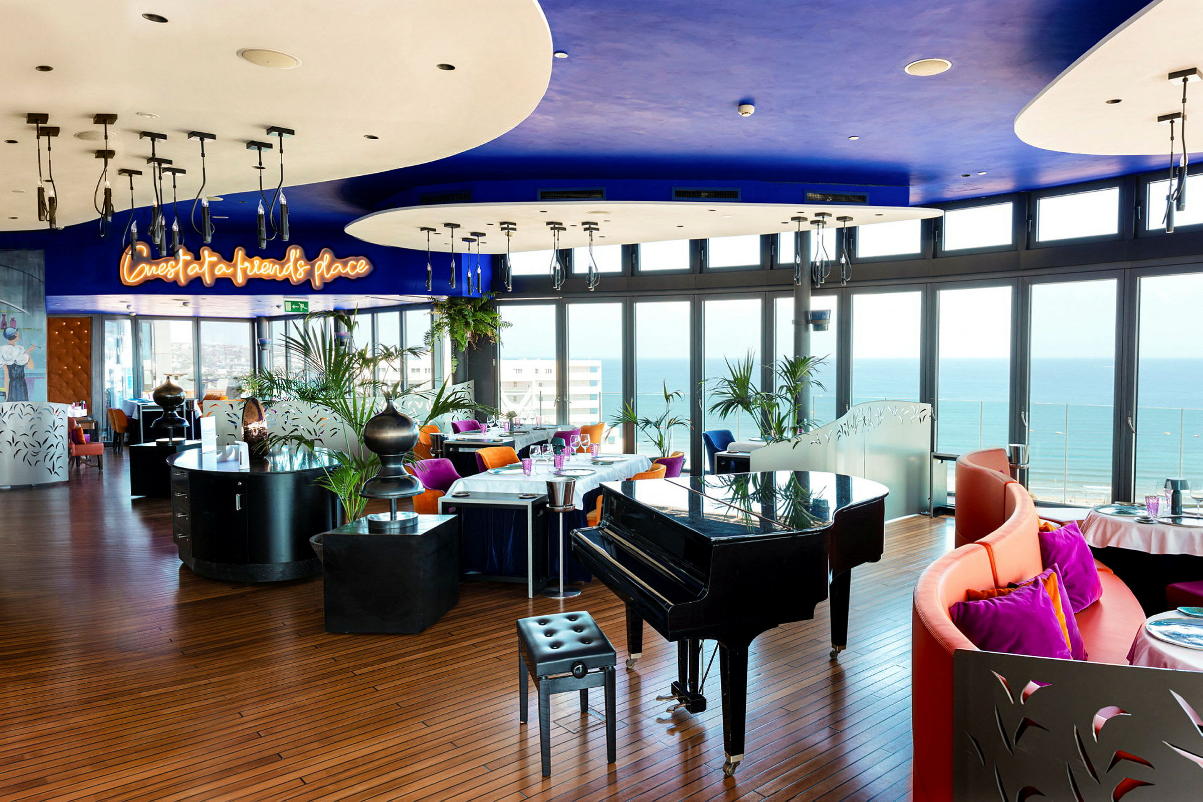 En restaurant med moderne innredning, fargerike stoler og et piano med utsikt over havet fra store vinduer. (AI-generert tekst)