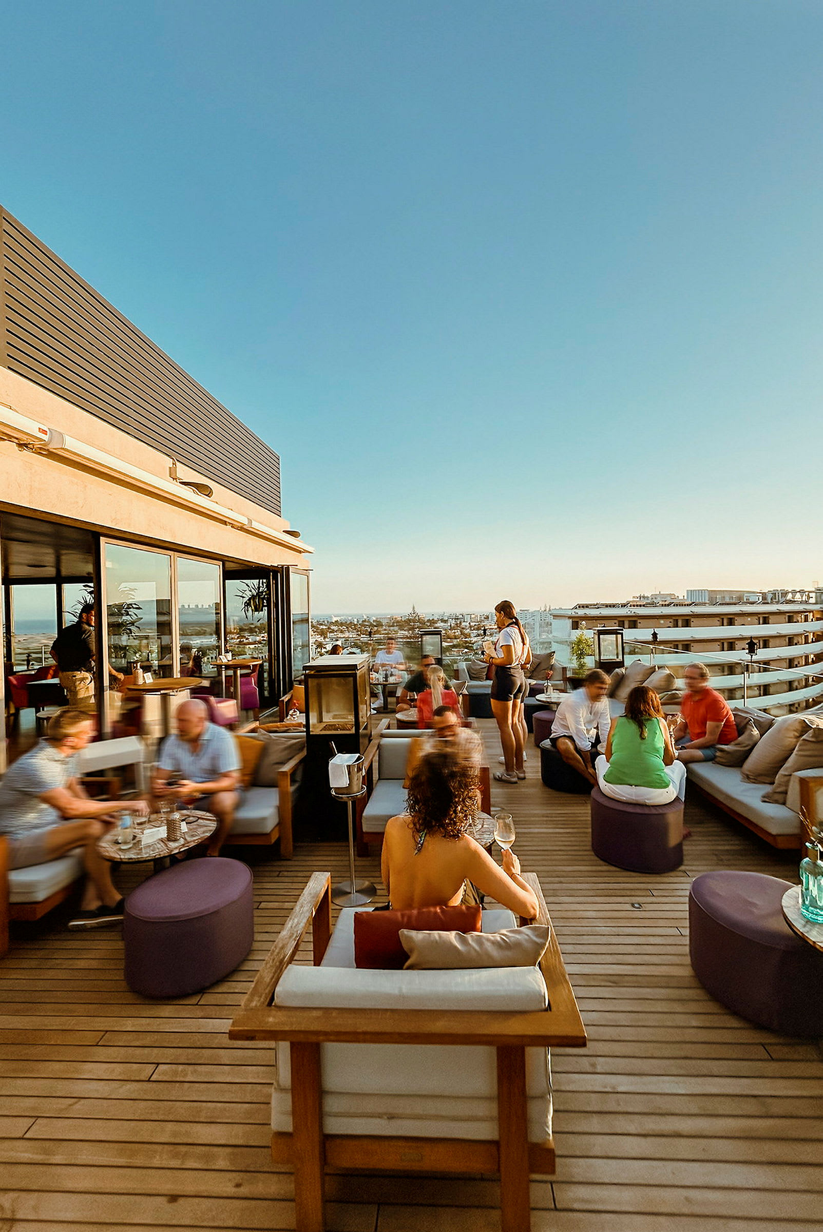 Utsikt over en takterrasse med sitteplasser og mennesker i en urban setting på en solrik dag. (AI-generert tekst)