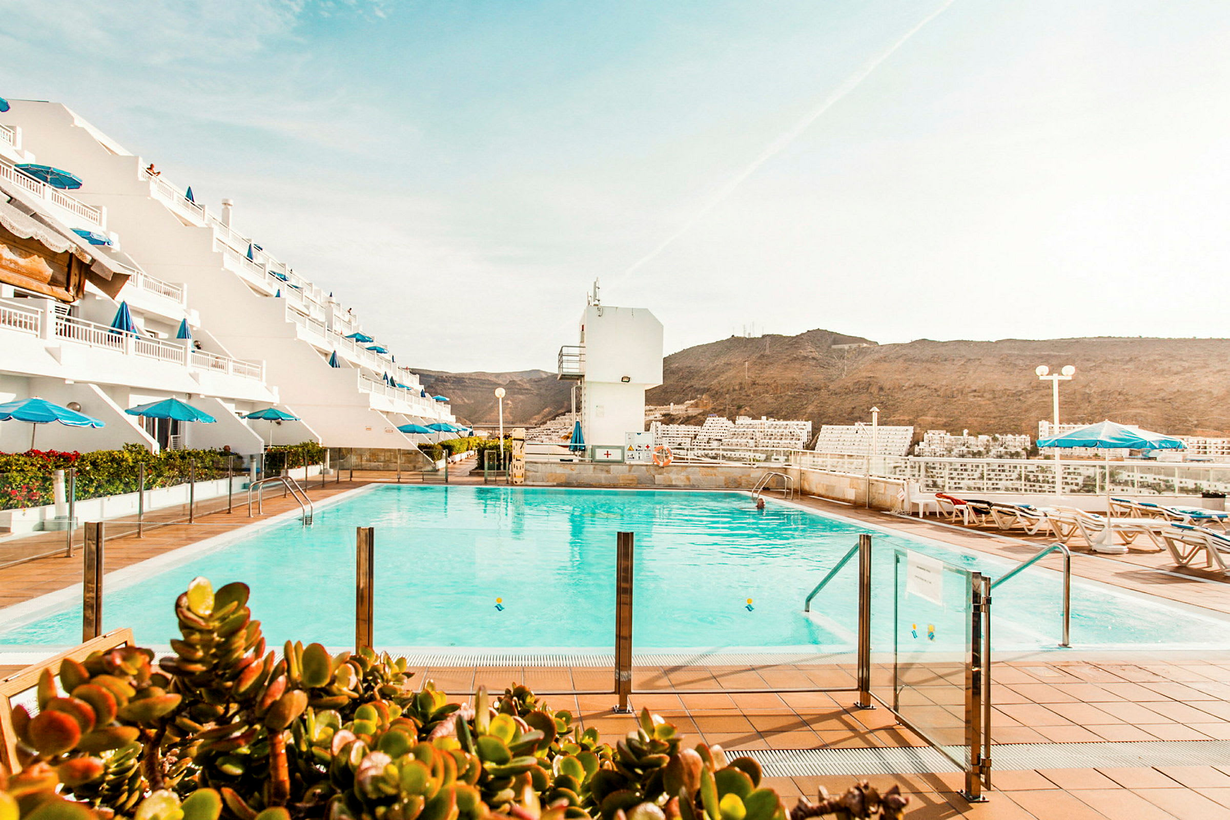 Bassengområde med solsenger og parasoller, omgitt av et hotell med hvite fasader og utsikt over fjellene. (AI-generert tekst)