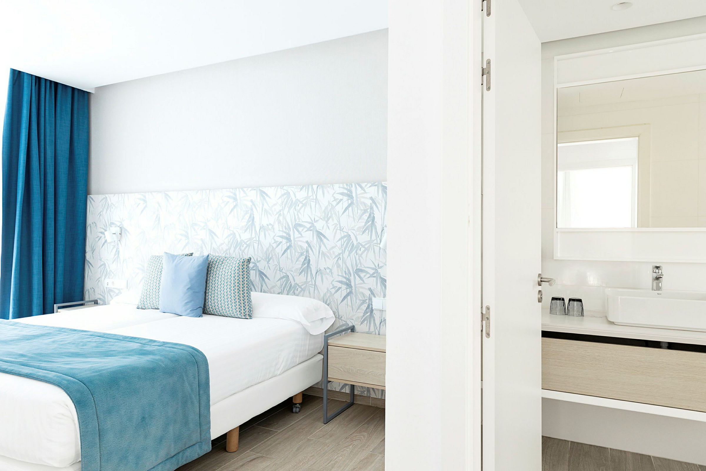 Et lyst hotellrom med en dobbeltseng med blå detaljer og et tilknyttet bad med synlig speil. (AI-generert tekst)