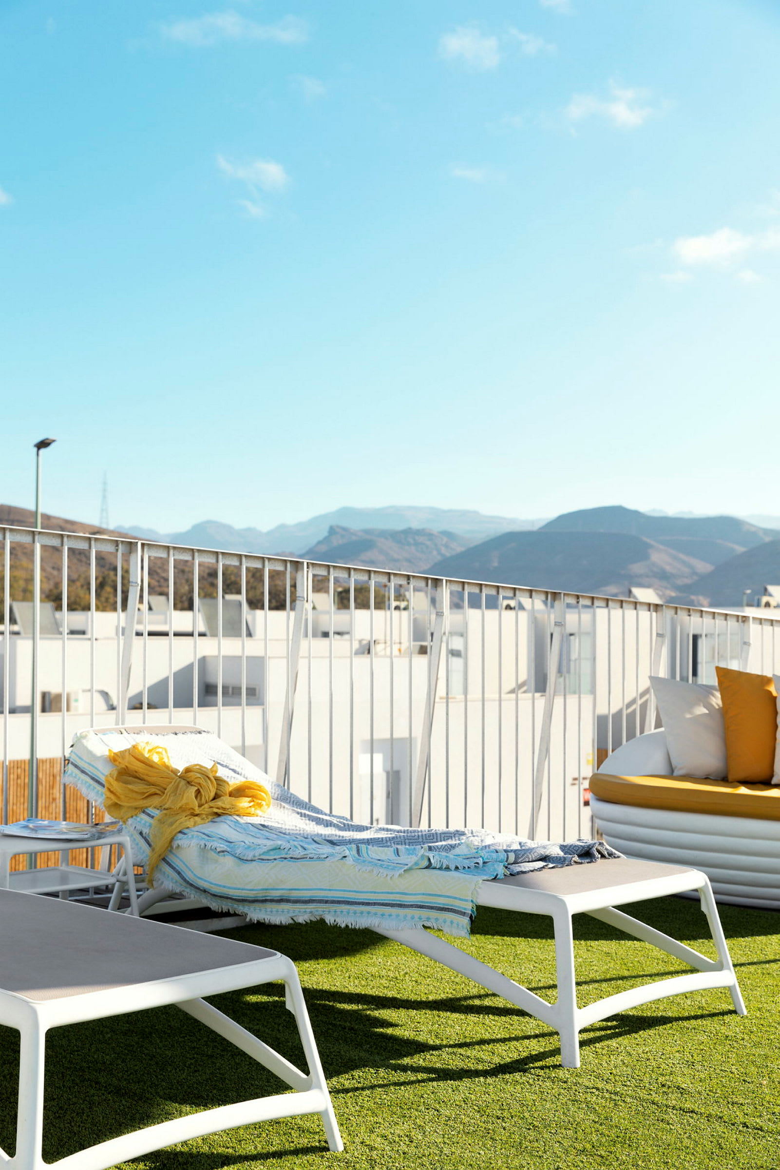 En solseng med et håndkle og et gult sjal ligger på en terrasse med kunstgress, med fjell og blå himmel i bakgrunnen. (AI-generert tekst)