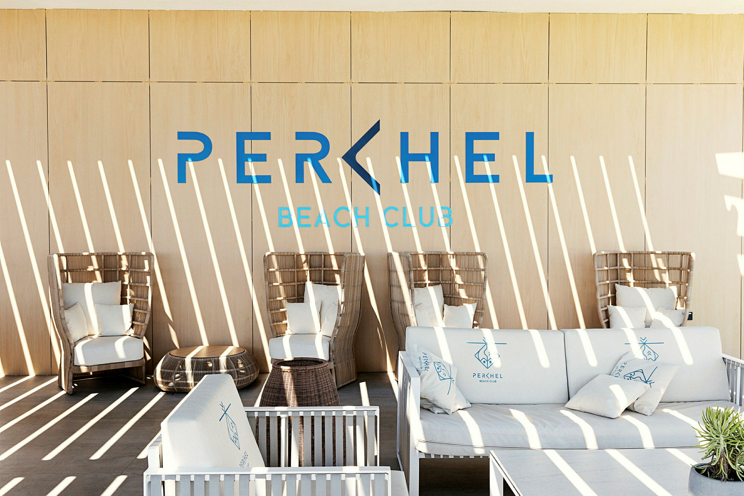 Et moderne loungeområde med kurvmøbler og sofaer med puter, samt teksten 'Perchel Beach Club' på veggen. (AI-generert tekst)