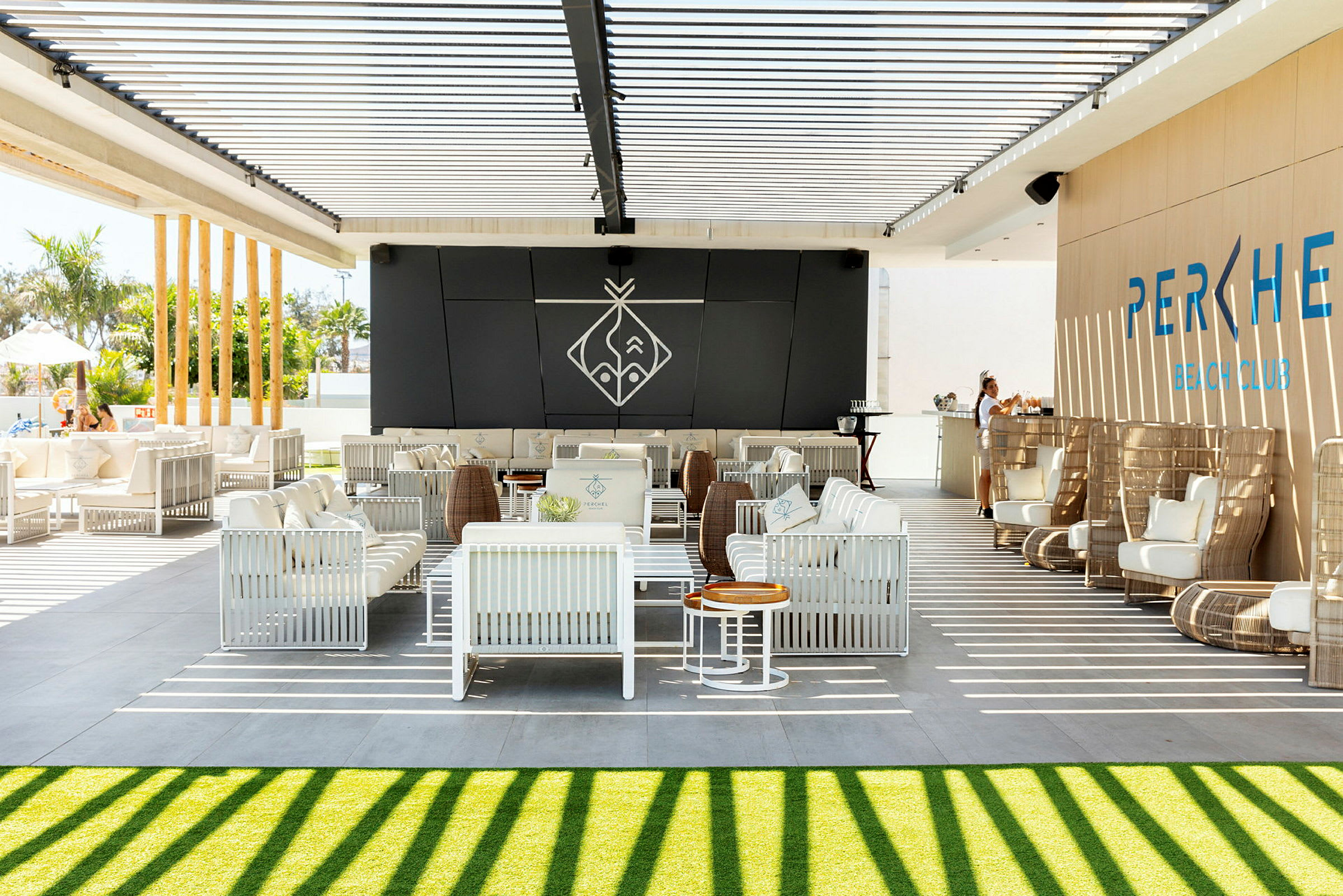 En utendørs lounge med hvite sittemøbler og en bar under en pergola med skyggelegging. (AI-generert tekst)