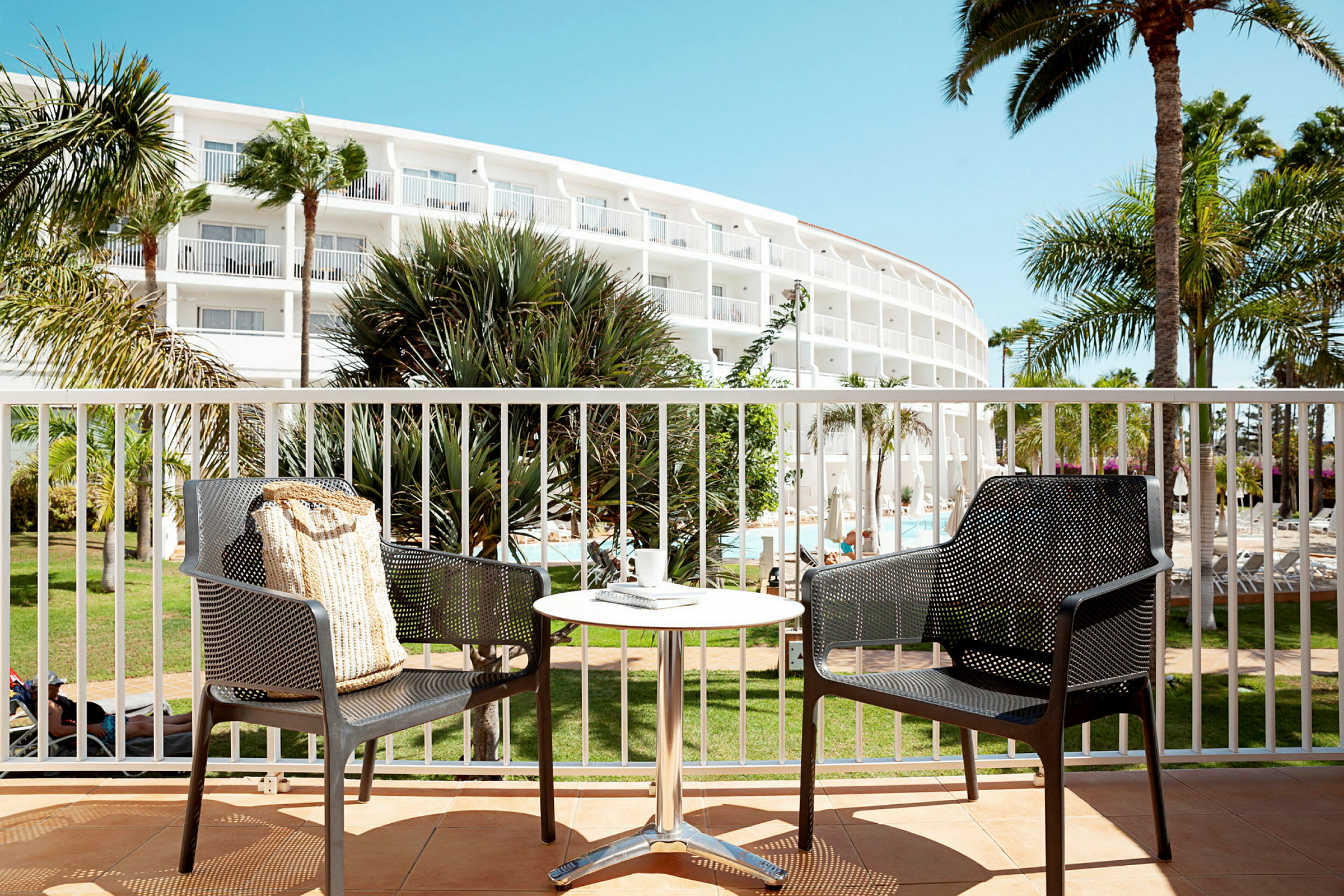 To stoler og et bord på en terrasse med utsikt over et hotellbygg og omliggende palmer. (AI-generert tekst)