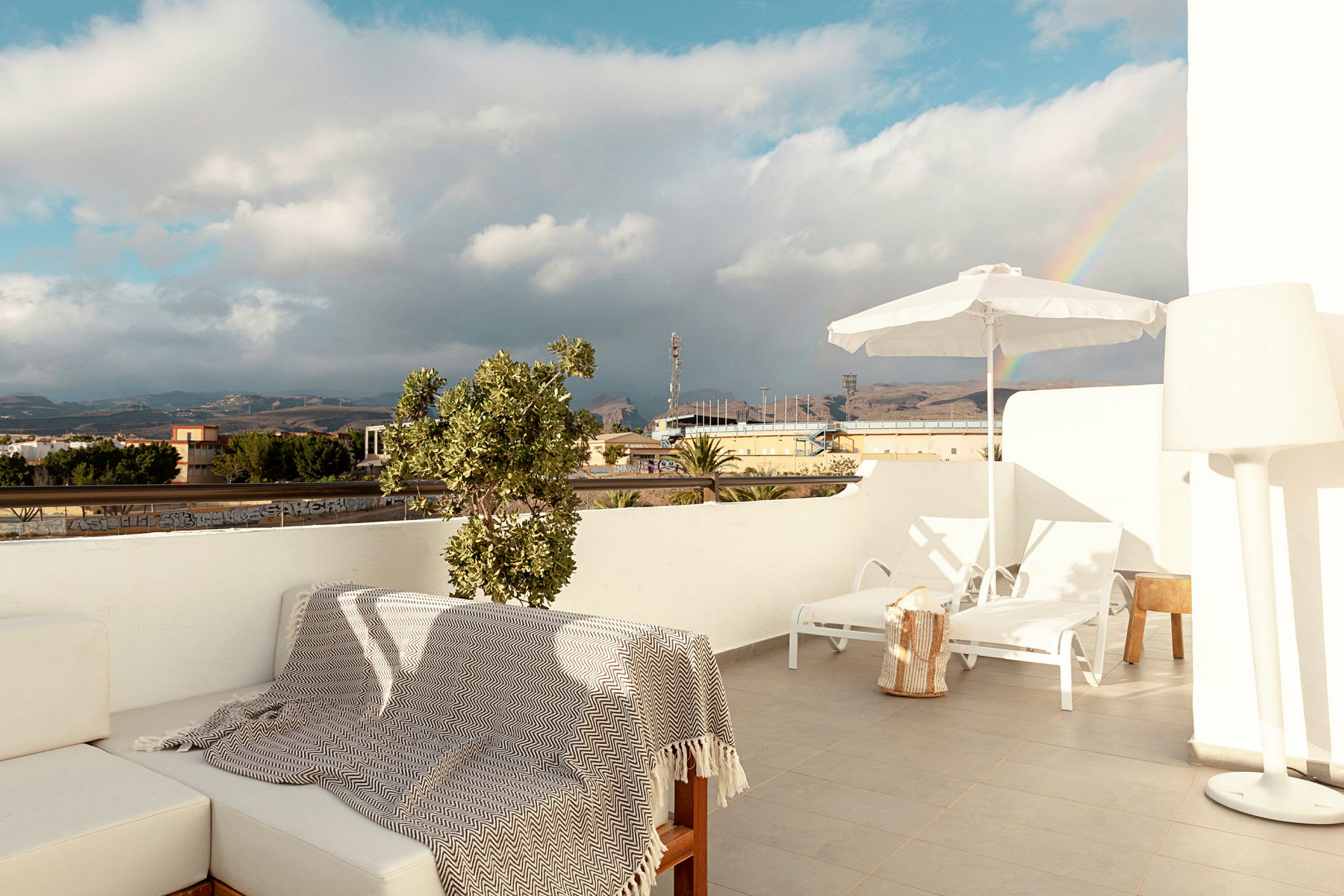 En terrasse med sittemøbler, solsenger og parasoll, med utsikt over et landskap med en regnbue i bakgrunnen. (AI-generert tekst)