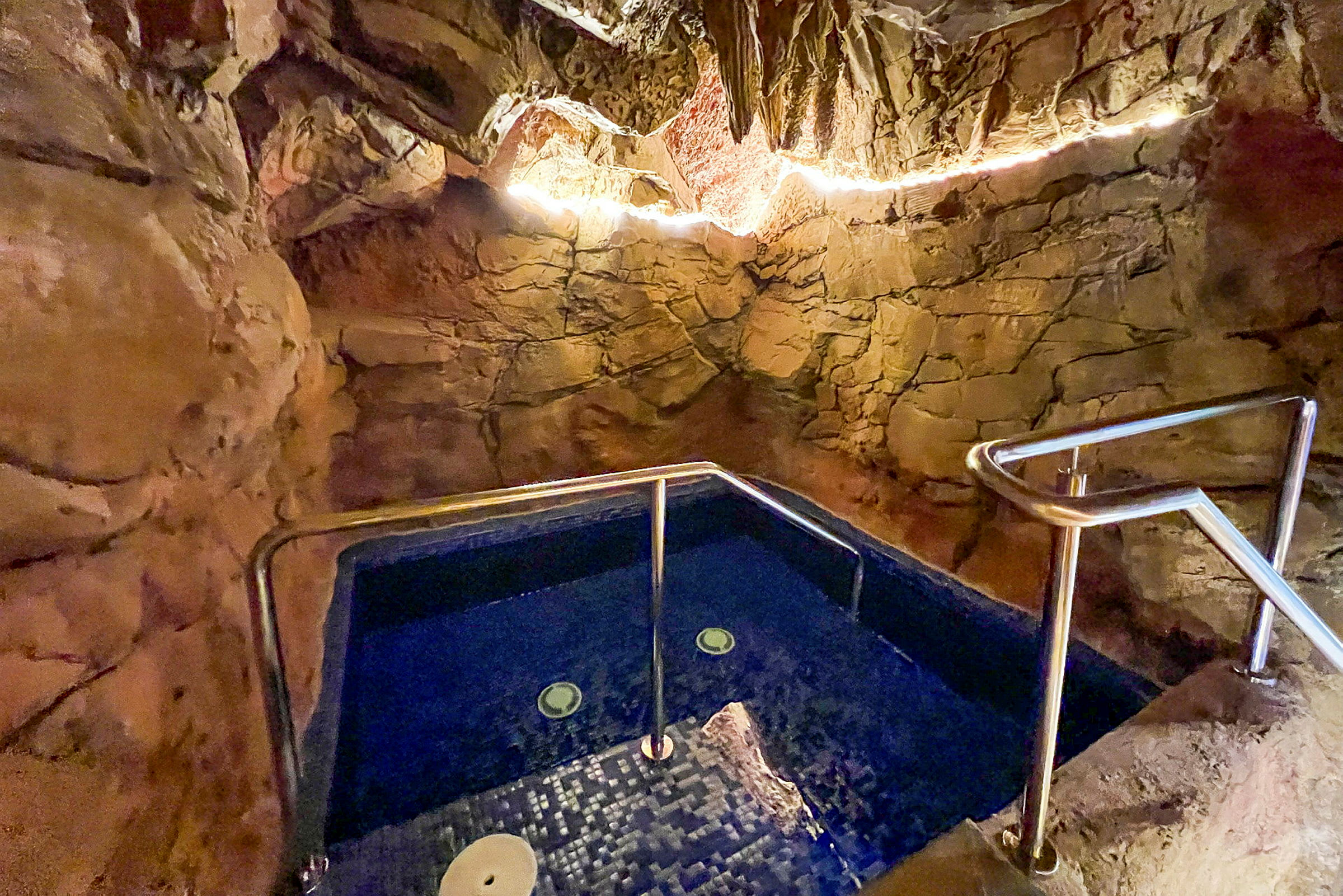 En jacuzzi omgitt av naturlignende steinvegger, med belysning langs taket. (AI-generert tekst)