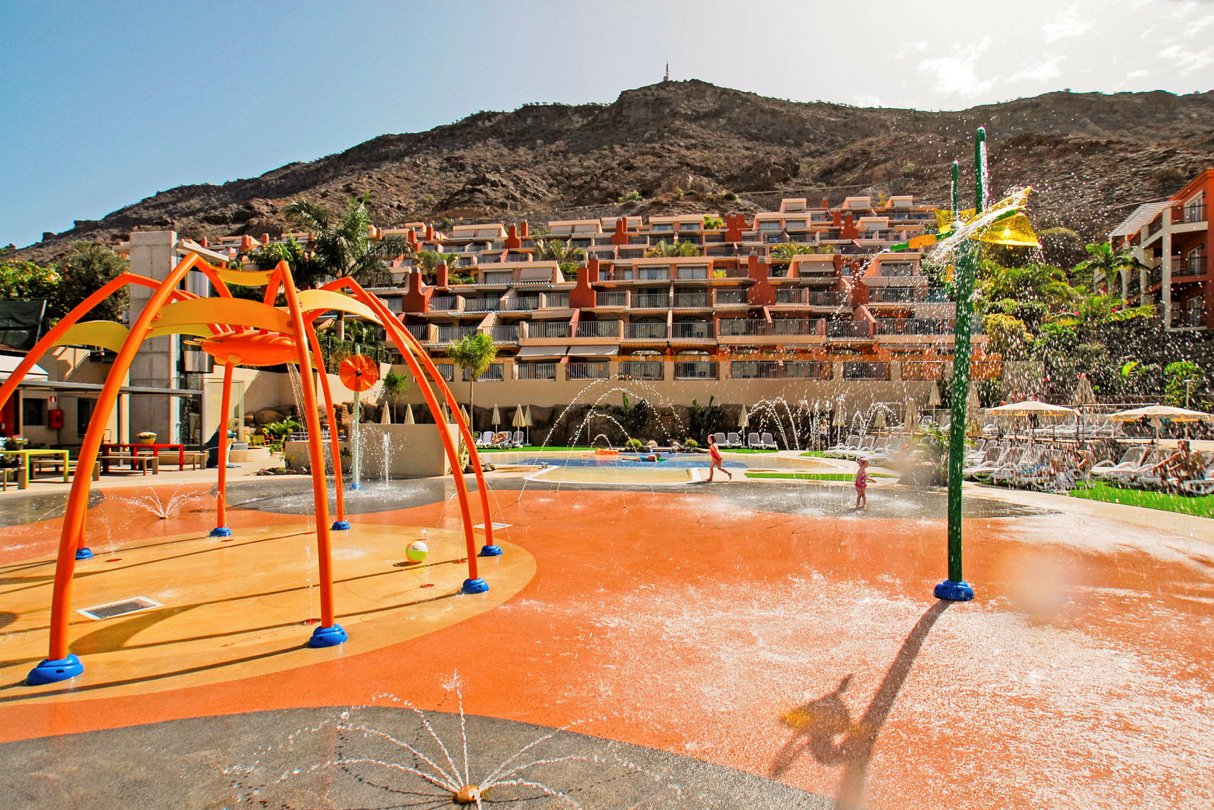 Lekeplass med vannfontener ved hotellkomplekset omgitt av fjell. (AI-generert tekst)