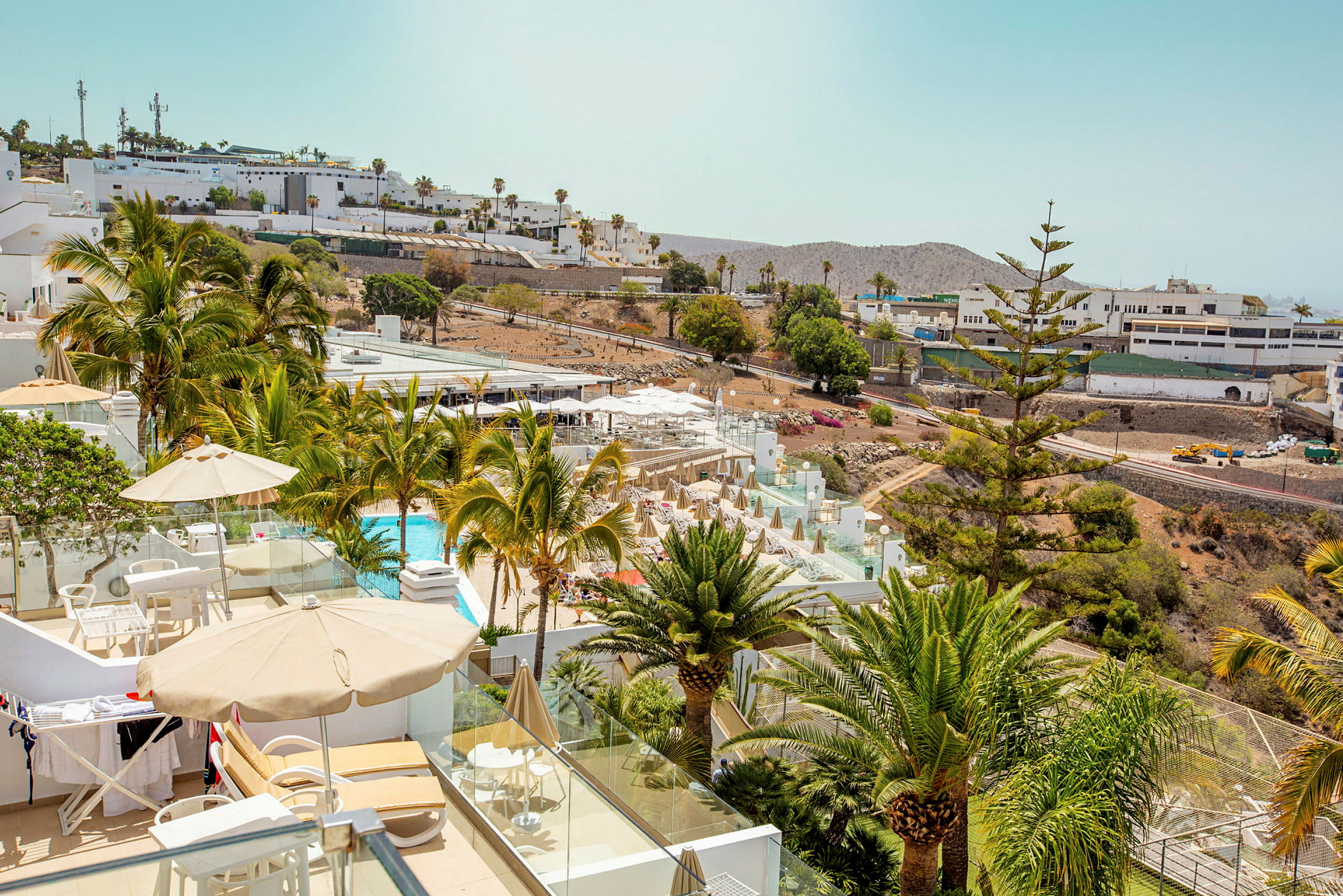 Utsikt over et hotellbasseng og terrasse med solsenger og parasoller, omgitt av grøntområder og palmer. (AI-generert tekst)